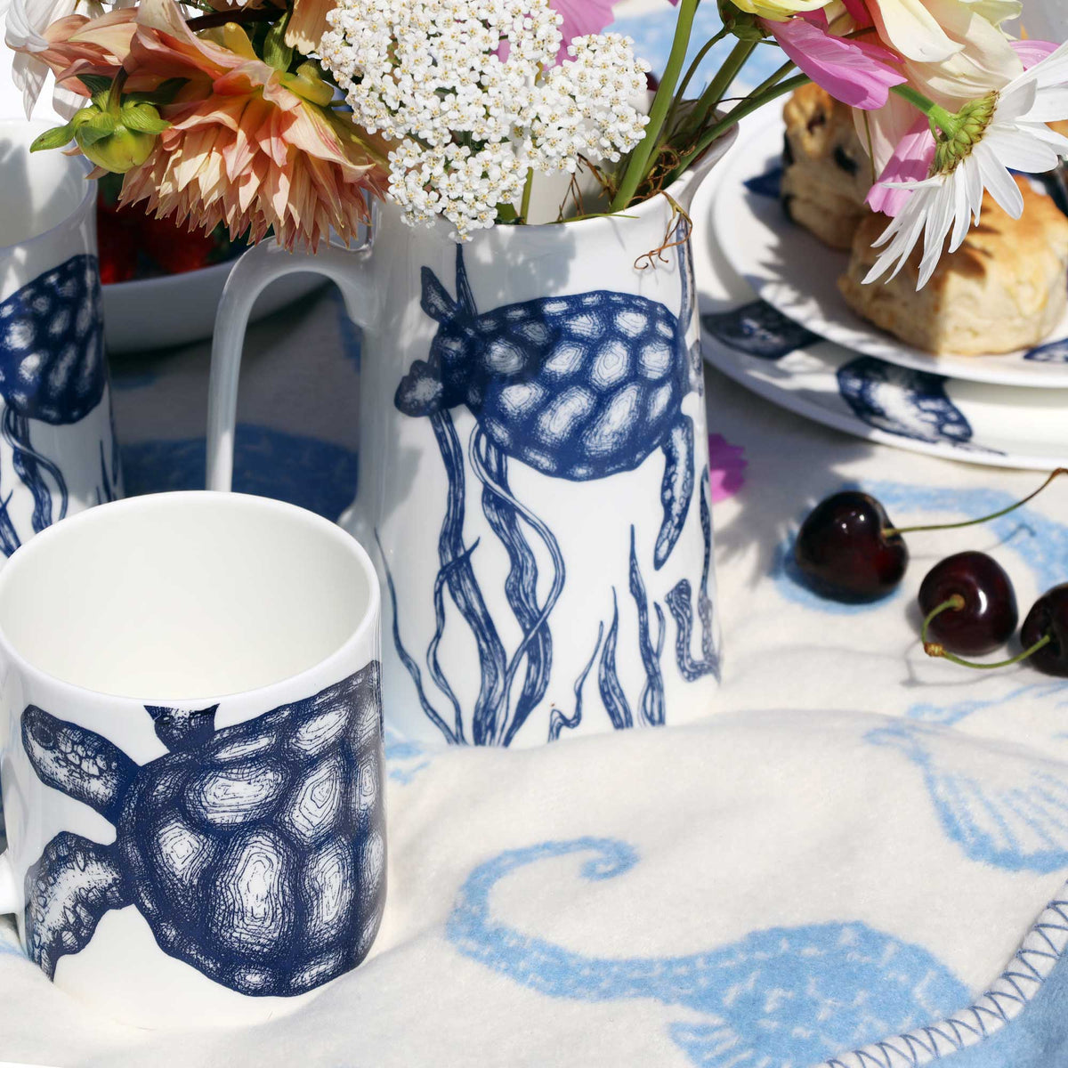 A white ceramic table setting with navy blue sea creature illustrations, including a turtle on a mug, a jellyfish on a pitcher, and a seahorse on a napkin. The scene is styled with flowers, pastries, and cherries on a white tablecloth.
