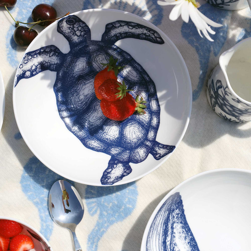 A white ceramic plate with a dark blue turtle illustration, styled on a patterned tablecloth with strawberries and a spoon nearby.