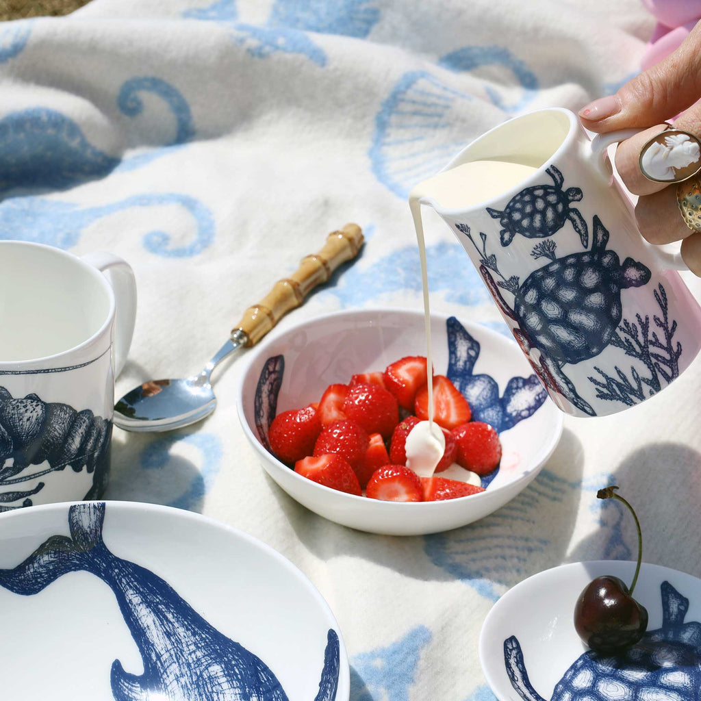 A hand pours milk from a blue-and-white ceramic pitcher into a bowl of sliced strawberries, with matching ceramic tableware and a patterned blanket in the background.