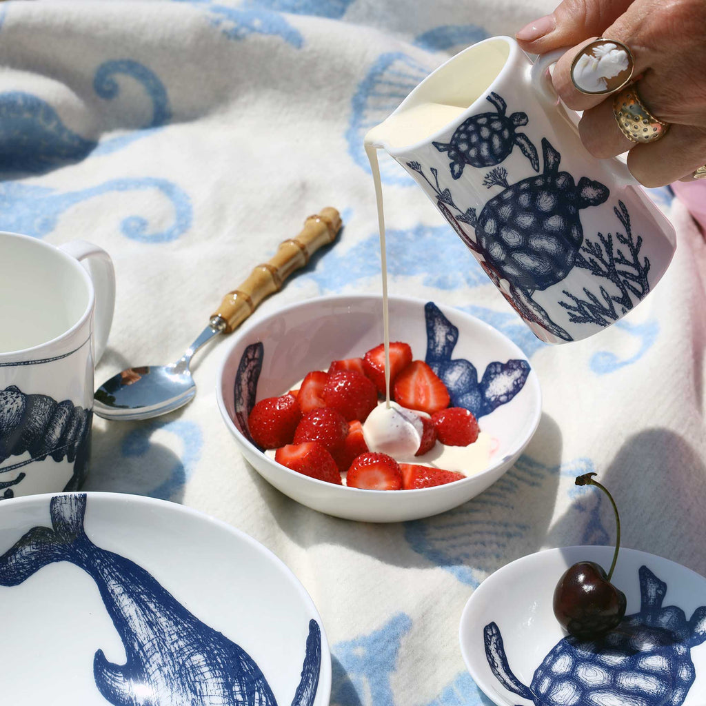 A hand pours milk from a white ceramic pitcher with a navy blue sea turtle design into a bowl of fresh strawberries. Other matching ceramic tableware with similar turtle illustrations is arranged on a blue and white patterned blanket.