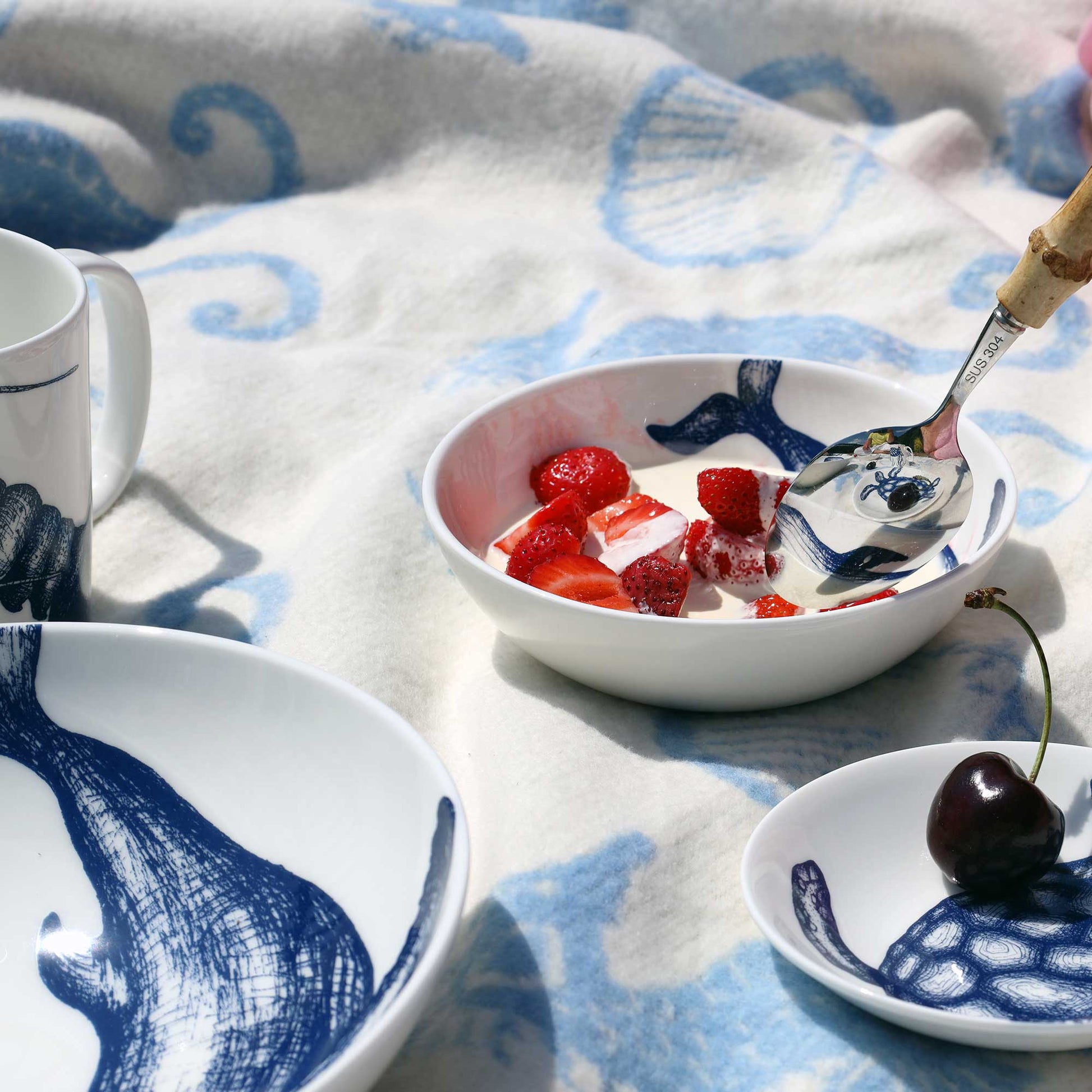 A styled tabletop scene with white ceramic dishes featuring navy blue abstract brushstroke designs, including a bowl with strawberries, a small plate with a cherry, and part of a mug. A patterned blanket is draped underneath on a light surface.