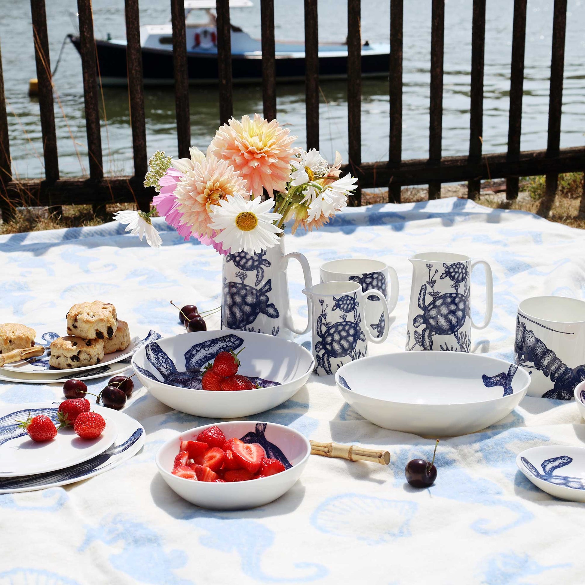 A styled outdoor table setting by the water with a white tablecloth printed in a light blue floral pattern. The table is decorated with a matching blue-and-white ceramic tableware set, including mugs, a pitcher, bowls, and a large serving dish, alongside food items like strawberries, cherries, and scones, and a vase of flowers in the center.