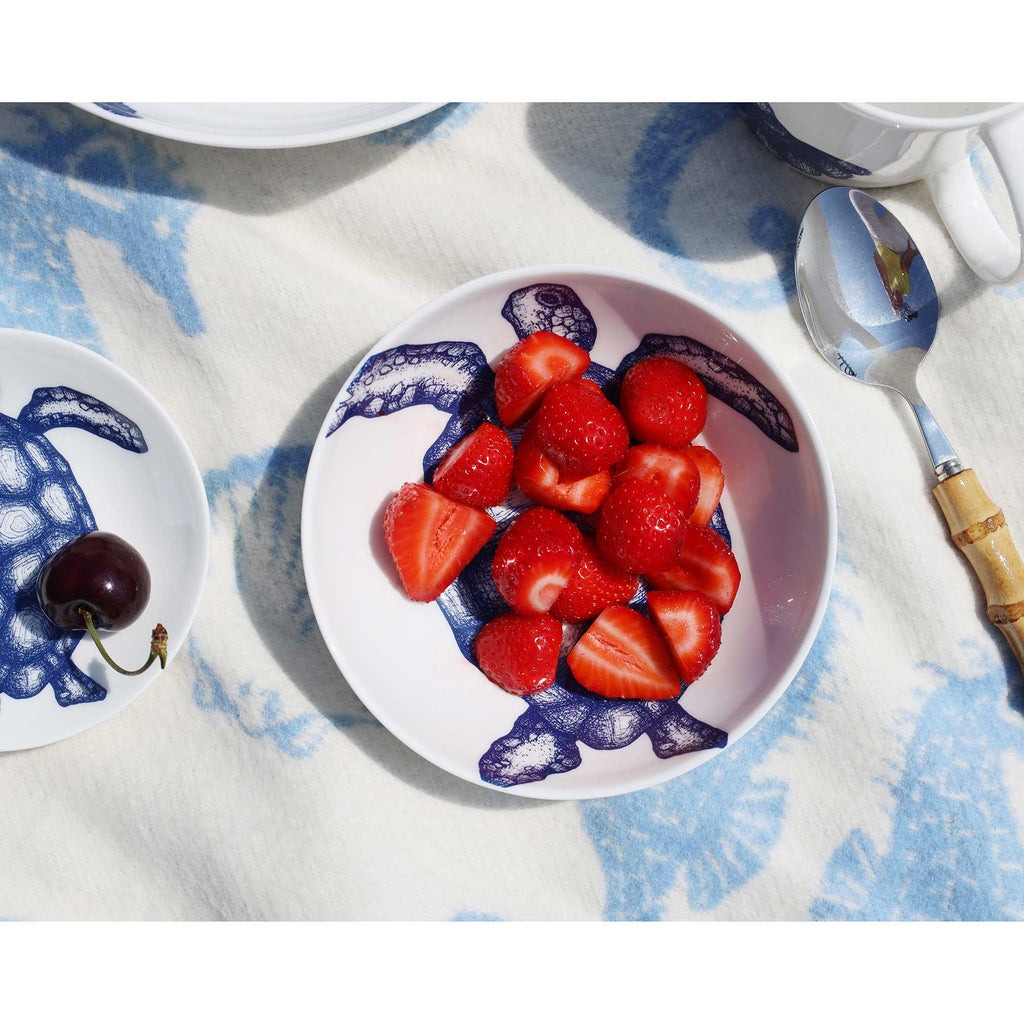 A white ceramic bowl with a blue turtle print, filled with sliced strawberries, placed on a blue and white patterned textile alongside a spoon, a cherry on a small plate, and other tableware.