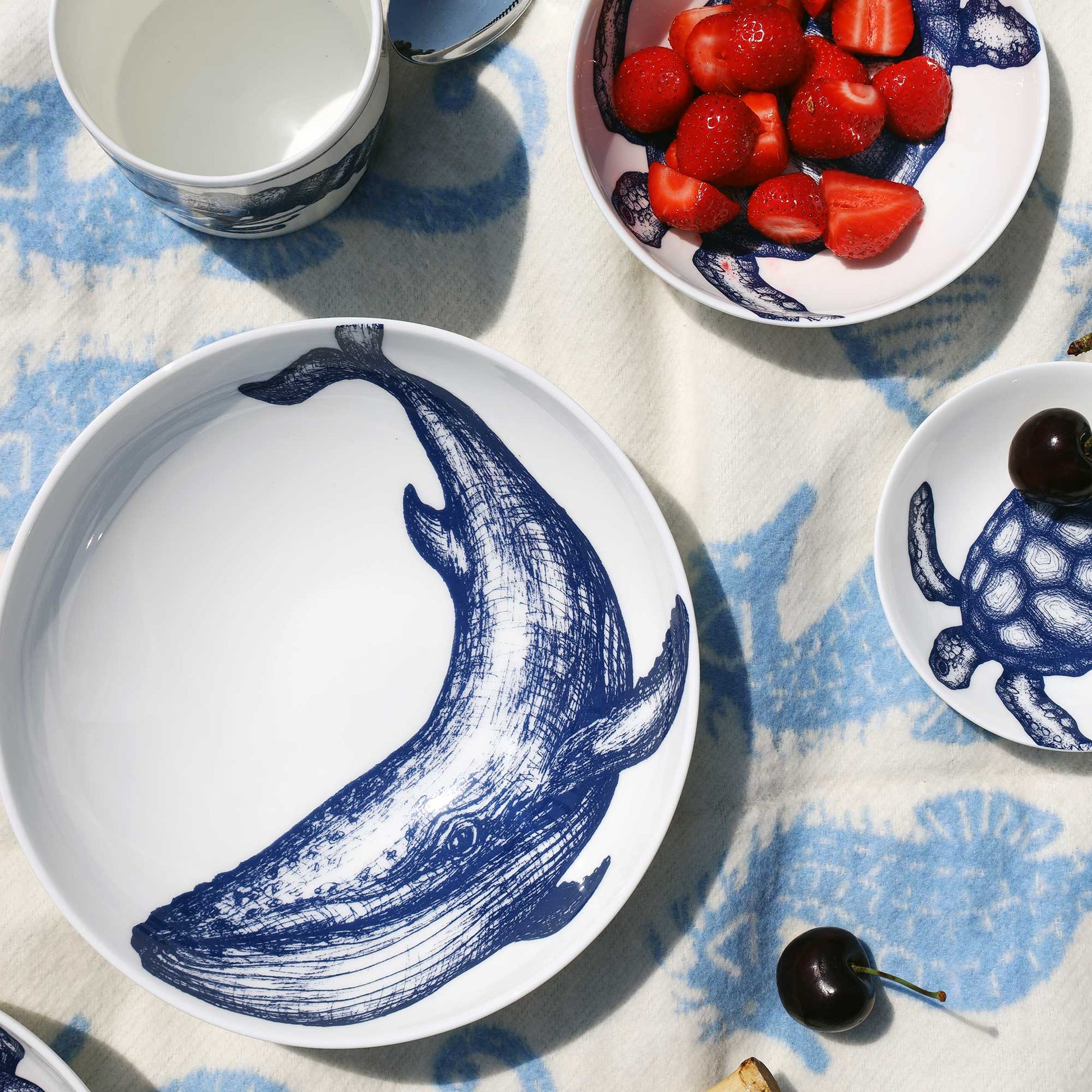 A white ceramic tableware set with a blue whale illustration, shown in a styled flat-lay on a blue and white patterned cloth. The set includes a large plate with a whale design, a small plate with a turtle design, a bowl of strawberries, and stacked cups.