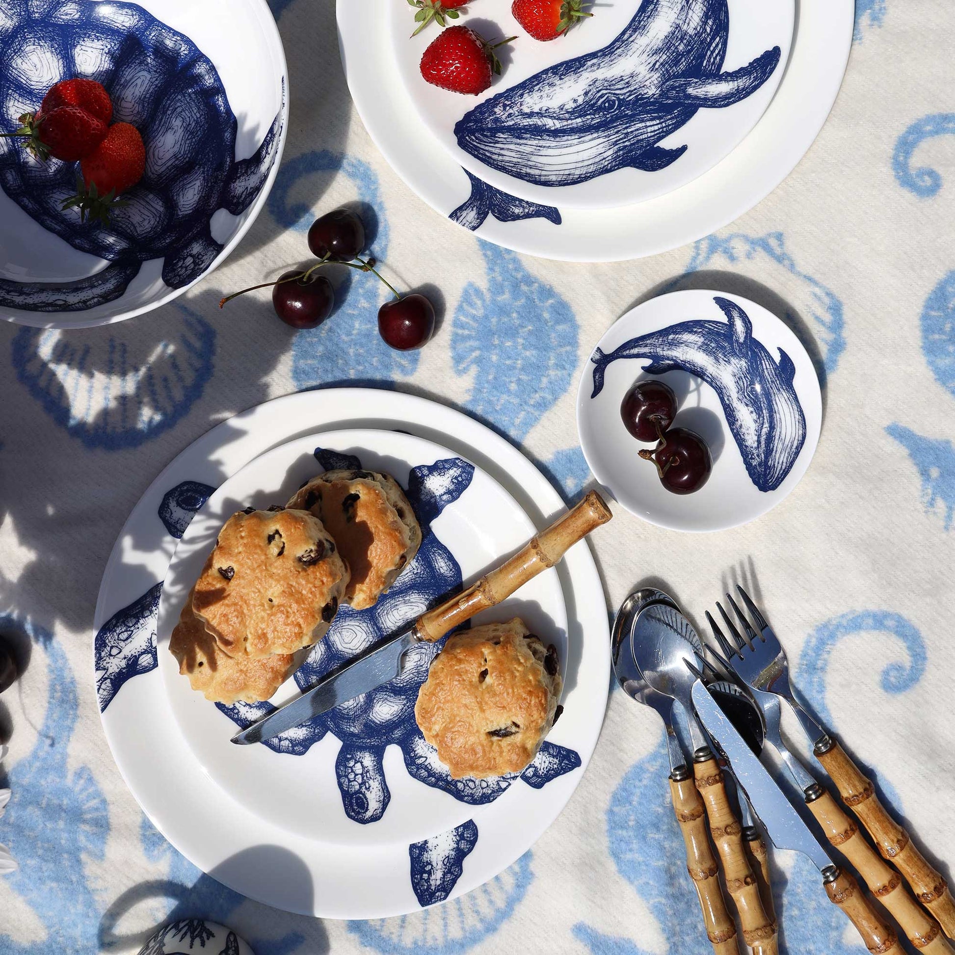 A styled overhead flat-lay of white ceramic plates with blue whale illustrations, set on a blue patterned tablecloth with strawberries, cherries, scones, a knife, and bamboo-handled cutlery.