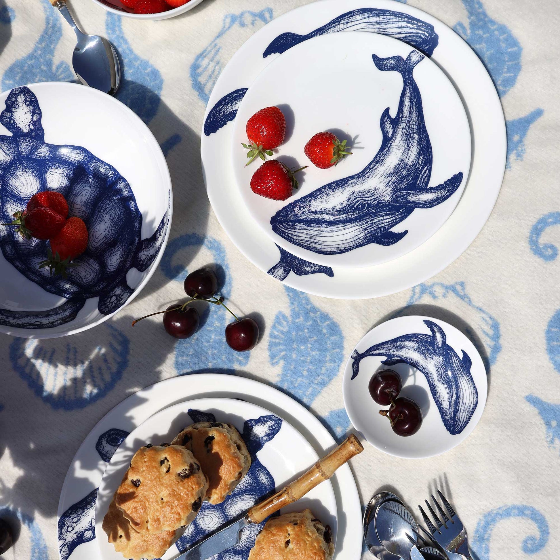 A styled overhead flat-lay of a white ceramic tableware set with navy blue whale illustrations, including plates, bowls, and a small dish, arranged on a blue patterned tablecloth with food props like strawberries, cherries, and scones.