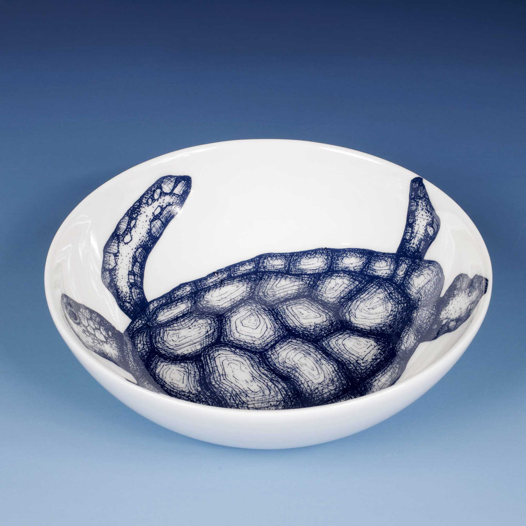 A white ceramic bowl featuring a detailed navy blue sketch of a sea turtle printed inside the rim, set against a blue gradient background.