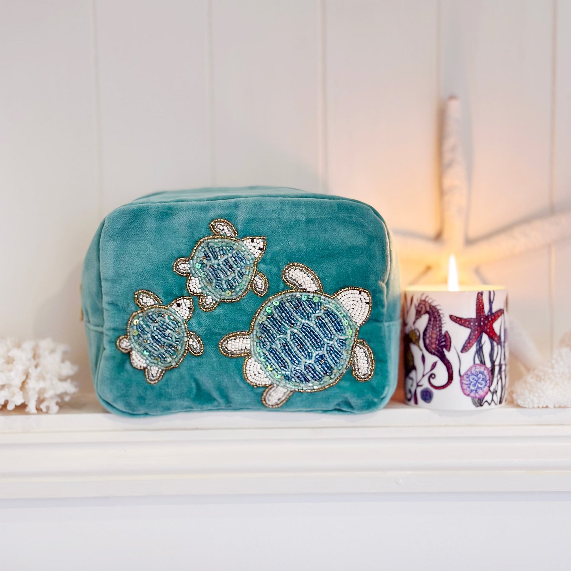 Teal velvet cosmetic bag featuring beaded embroidery of three sea turtles, styled on a white shelf with a lit coastal-themed candle, coral decor, and a white starfish against a paneled background, creating a serene seaside ambiance.