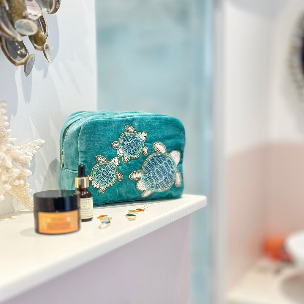 Teal velvet cosmetic bag adorned with intricate beaded sea turtle embroidery, displayed on a bathroom shelf alongside a starfish, skincare products, and small trinkets, with soft natural light enhancing the coastal-inspired setting.