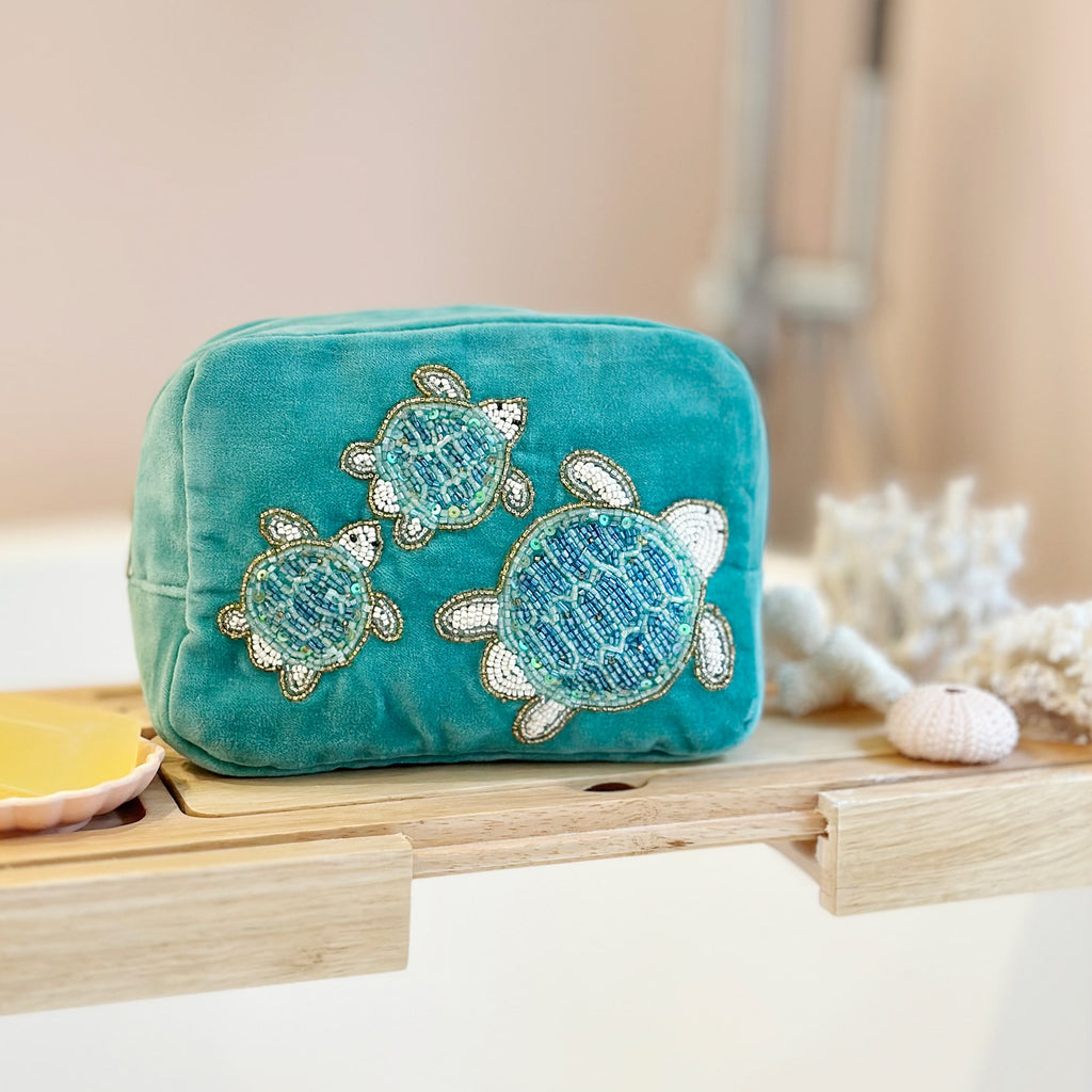 Teal velvet cosmetic bag decorated with intricate beaded embroidery of three sea turtles, displayed on a wooden bath tray beside decorative coral, a soap dish, and a pink sea urchin, creating a relaxing, ocean-inspired bathroom scene.