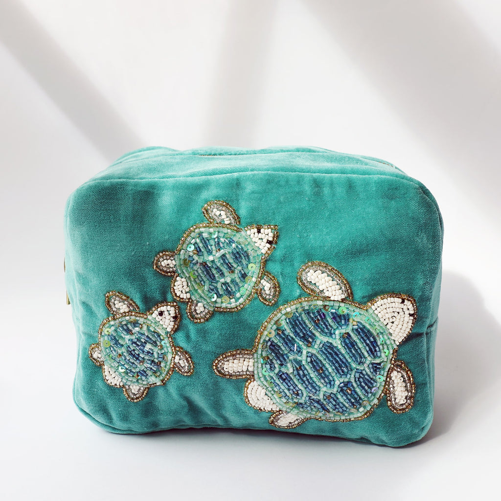 A turquoise velvet cosmetic bag adorned with beaded embroidery of three sea turtles in different sizes. The turtles are crafted from shimmering blue, green, white, and gold beads, creating a textured and eye-catching design. The bag is photographed in soft natural light against a neutral background, casting gentle shadows.