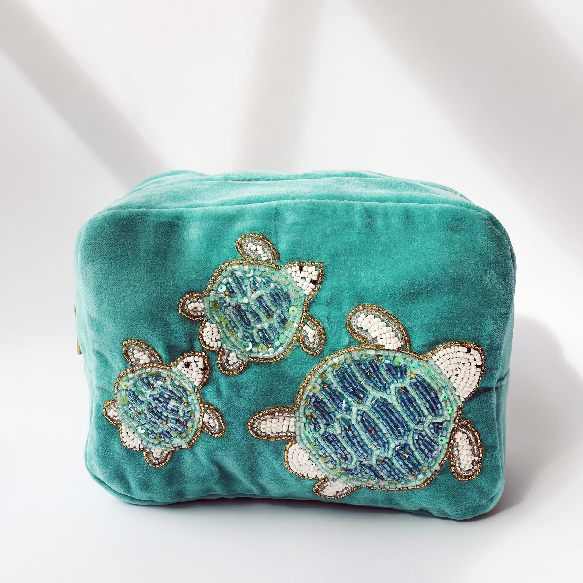 A turquoise velvet cosmetic bag adorned with beaded embroidery of three sea turtles in different sizes. The turtles are crafted from shimmering blue, green, white, and gold beads, creating a textured and eye-catching design. The bag is photographed in soft natural light against a neutral background, casting gentle shadows.