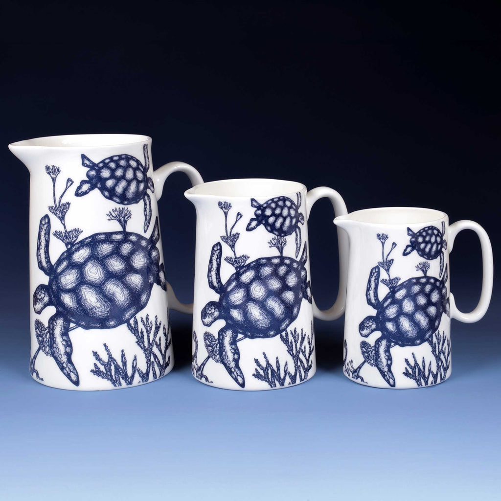 Three white ceramic pitchers of different sizes with blue sea turtle and coral patterns, arranged against a dark blue gradient background.