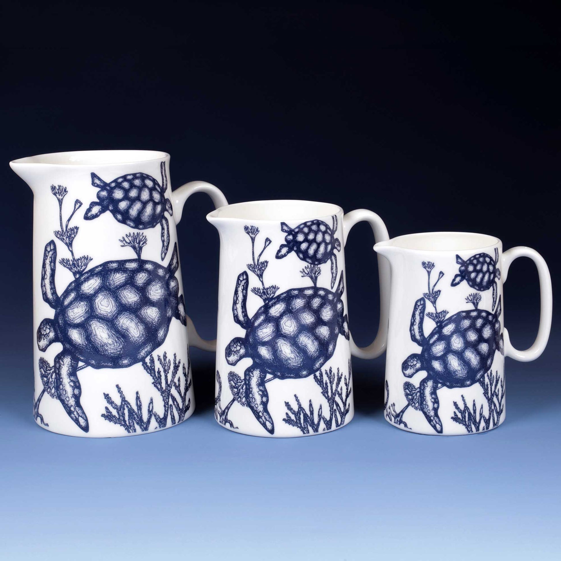 Three white ceramic pitchers of different sizes with blue sea turtle and coral patterns, arranged against a dark blue gradient background.