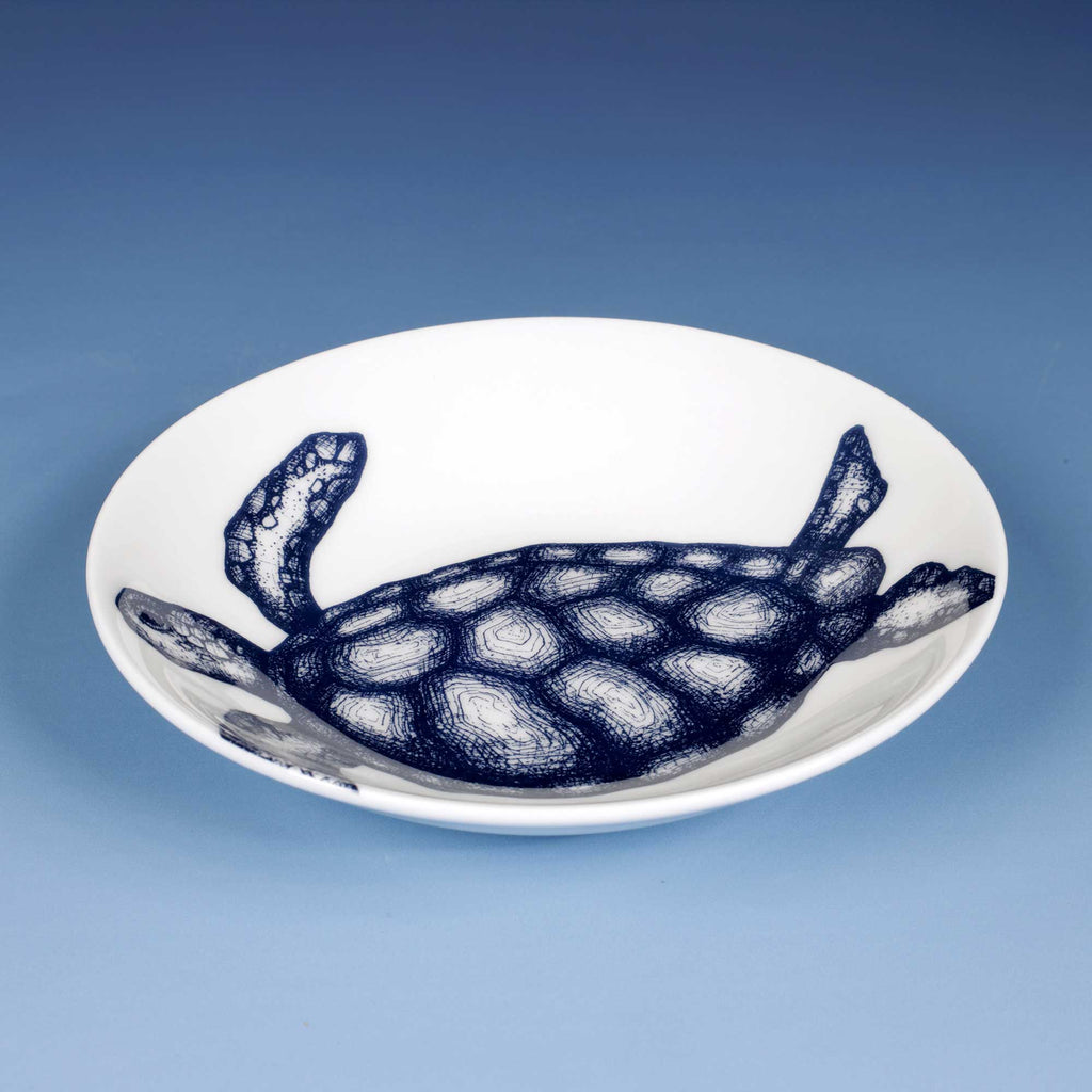 A white ceramic shallow bowl featuring a detailed navy blue sketch of a sea turtle, set against a smooth blue gradient background.
