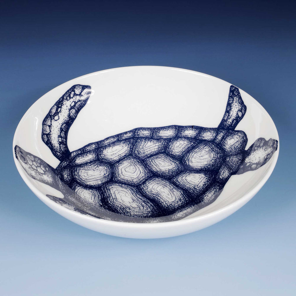 A white ceramic bowl featuring a detailed navy blue sketch of a sea turtle, set against a blue gradient background.