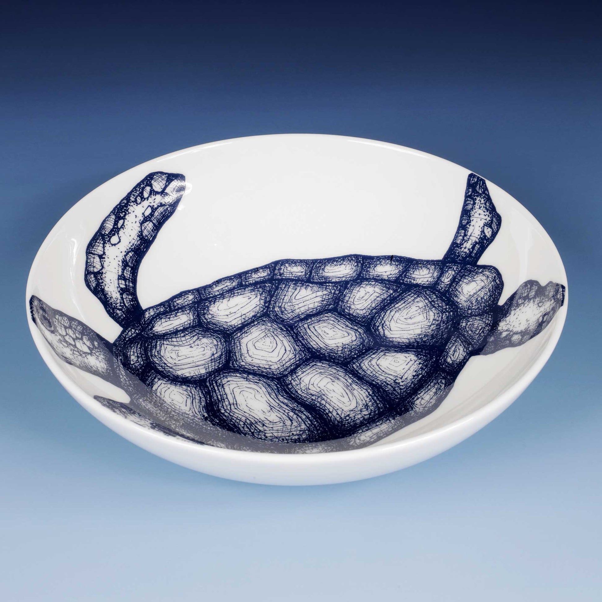 A white ceramic bowl featuring a detailed navy blue sketch of a sea turtle, set against a blue gradient background.