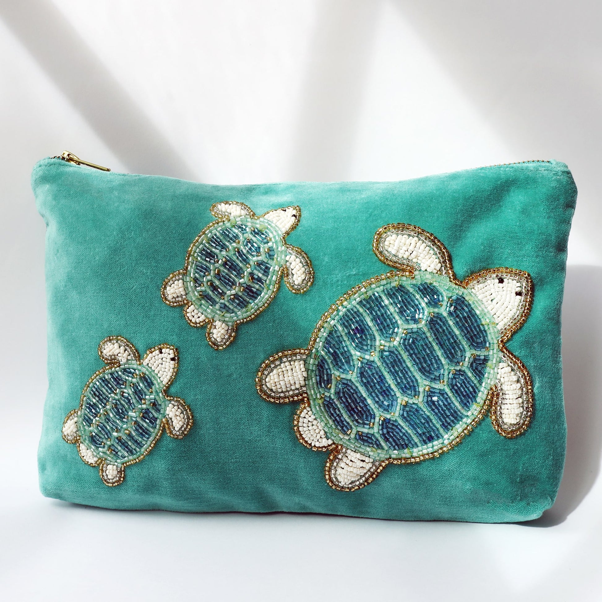 A turquoise velvet pouch featuring intricate beaded embroidery of three sea turtles in shades of blue, white, and gold. The pouch has a gold zip closure and is photographed in natural lighting with soft shadows, accentuating the vibrant colors and texture of the beadwork.
