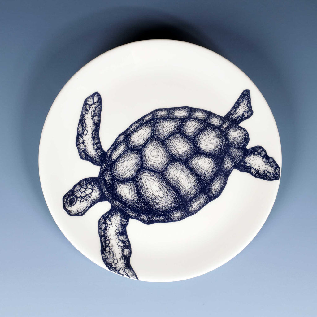 A round white plate featuring a detailed navy blue line art illustration of a sea turtle, shown from a top-down view against a soft blue gradient background.