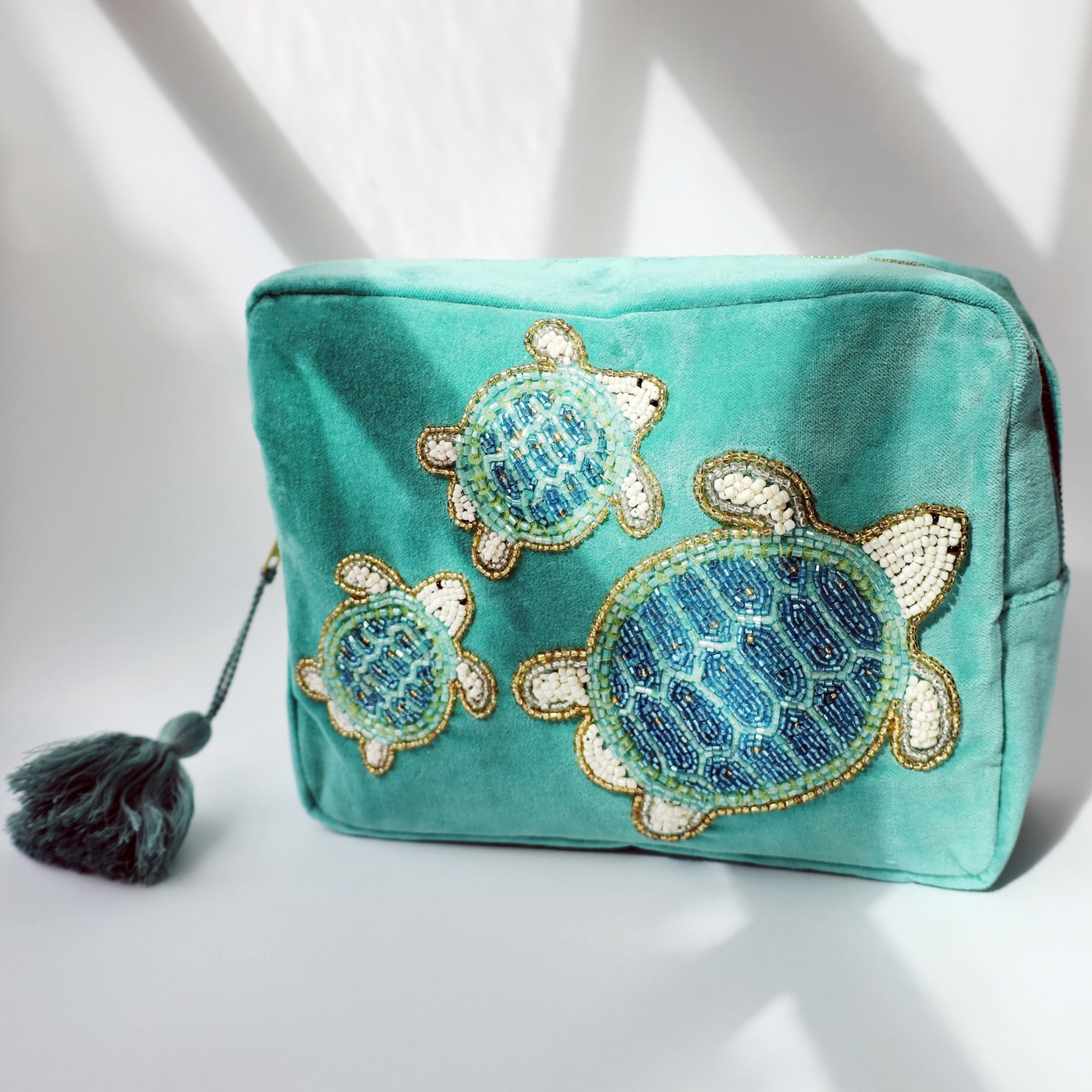 A turquoise velvet washbag adorned with beaded embroidery of three sea turtles in shimmering shades of blue, white, and gold. The washbag features a matching teal tassel zip pull and is photographed in soft, natural lighting with dramatic shadows, highlighting the texture and sparkle of the beadwork.