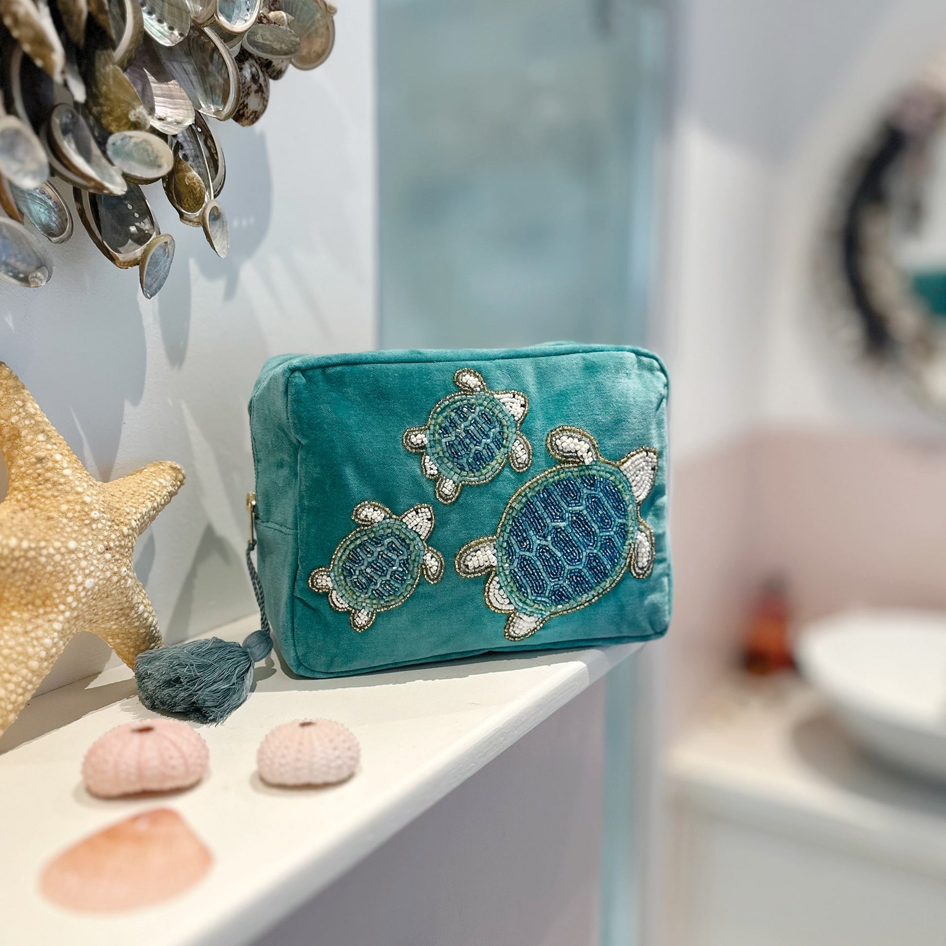 Turquoise velvet cosmetic bag featuring three beaded sea turtles, displayed on a bathroom shelf with a starfish, face cream, serum dropper bottle, and delicate shell earrings, creating a serene coastal-themed setting.