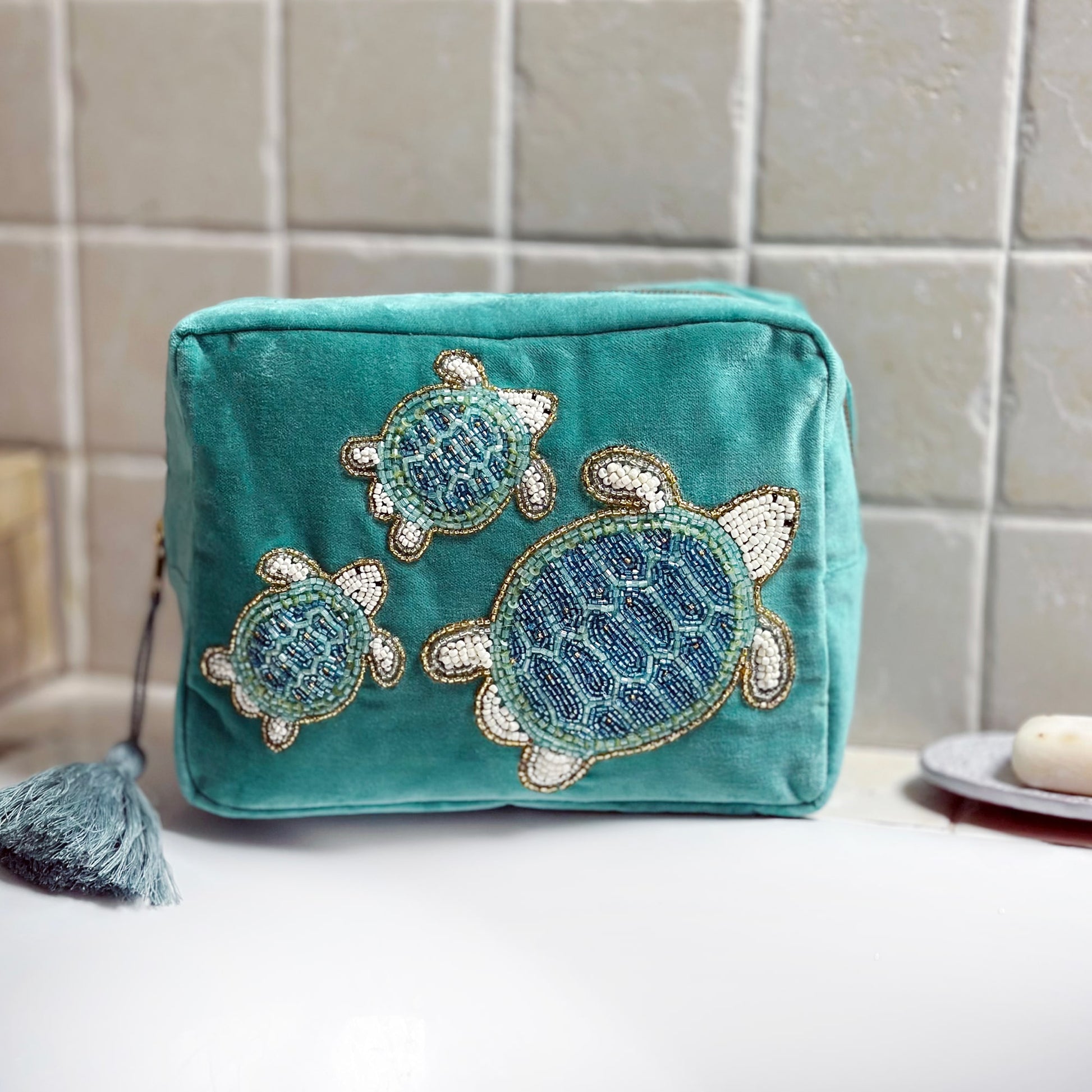 Turquoise velvet wash bag adorned with three intricately beaded sea turtles, placed on the edge of a white bathtub with a tiled wall in the background and a soap dish to the side, creating a calming, coastal bathroom aesthetic.