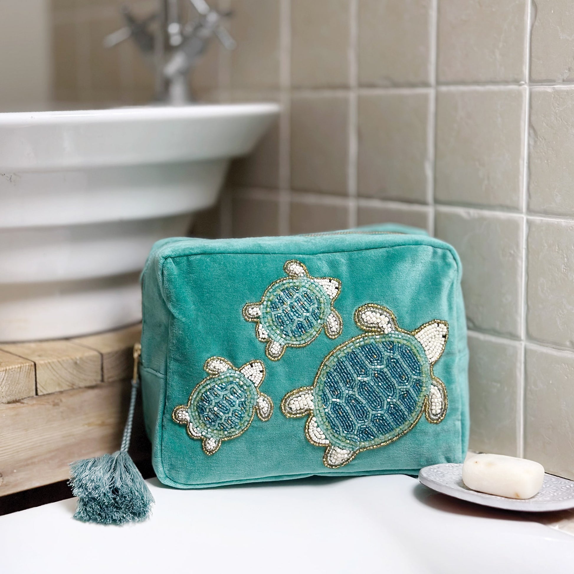 Turquoise velvet washbag adorned with three beaded sea turtles, styled on a bathroom counter next to a white ceramic sink, rustic wood accents, and a soap dish, creating a coastal and spa-like atmosphere.