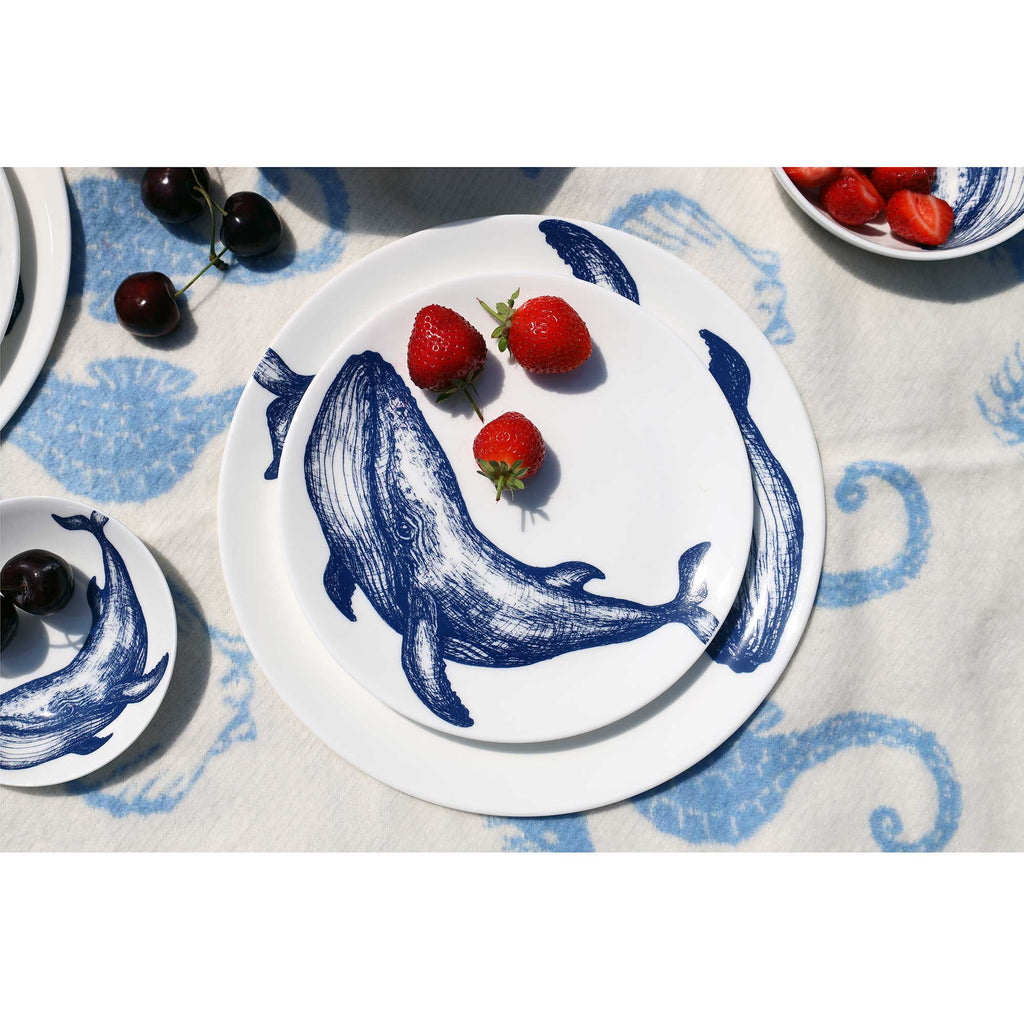 A stack of white ceramic plates with blue whale illustrations, styled on a blue and white patterned tablecloth with strawberries and cherries.