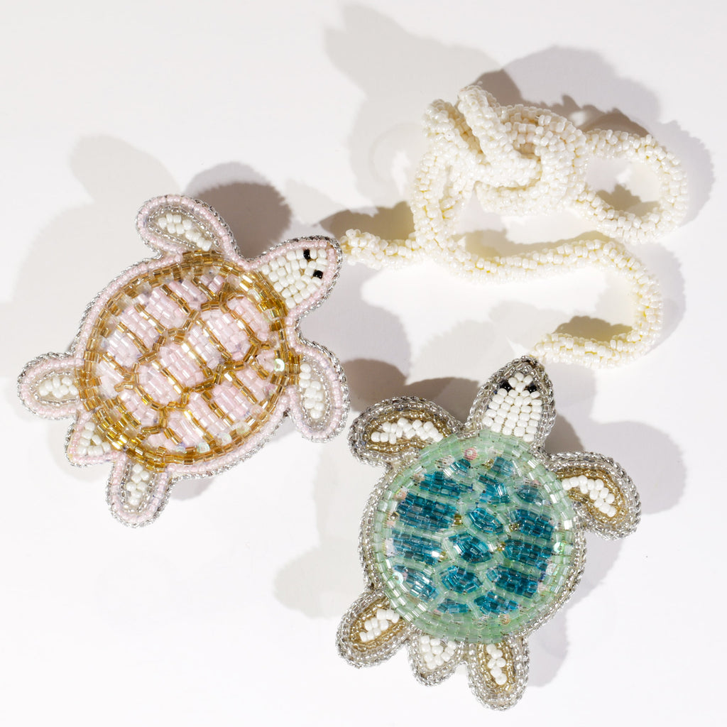Two beaded turtle bag charms, one in gold and pink tones and the other in turquoise and silver, lying on a white surface with ivory beaded loops attached.