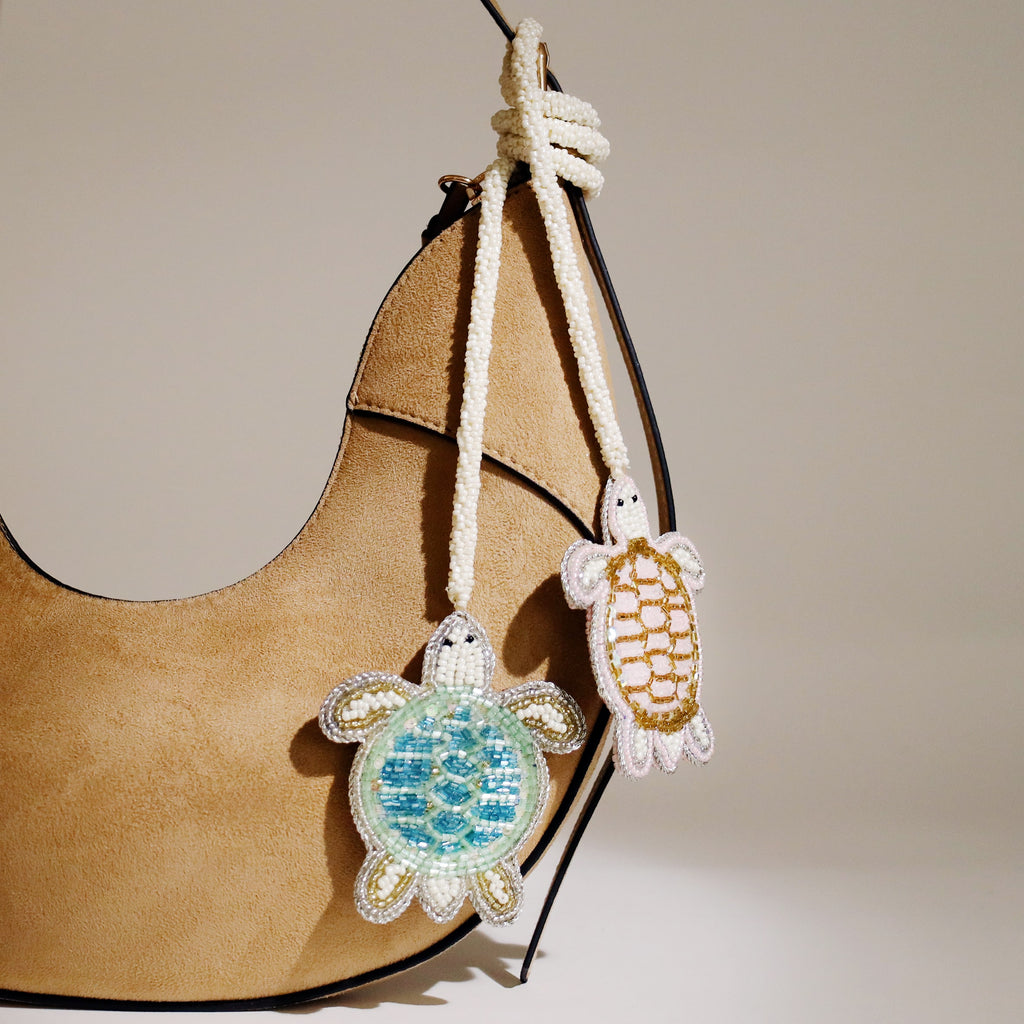 Two beaded turtle bag charms, one in blue and one in gold tones, hanging from the handle of a tan suede handbag.