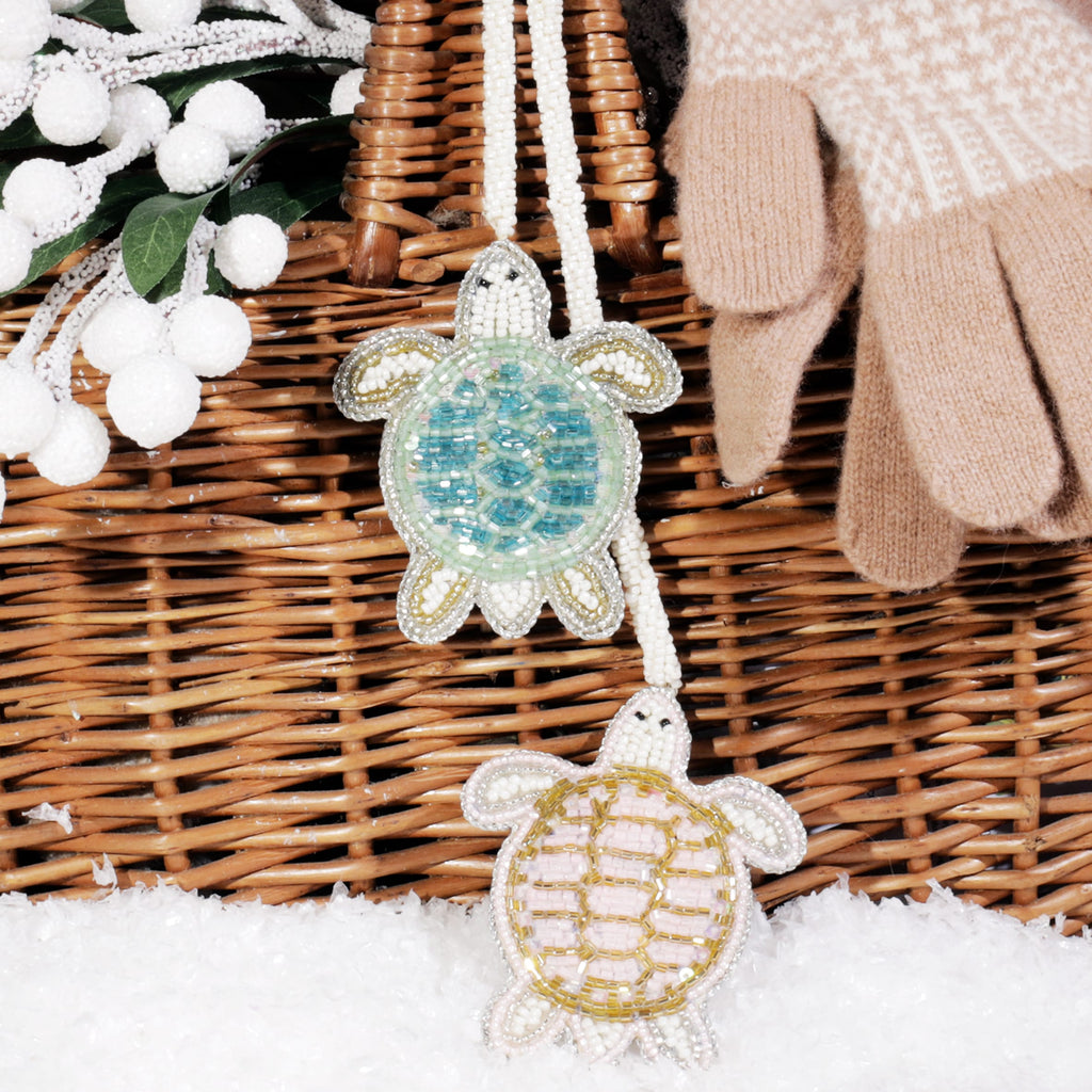 Two beaded turtle bag charms, one turquoise and one gold, hanging from a wicker basket beside a pair of beige wool gloves and frosted white berries on a snowy surface.
