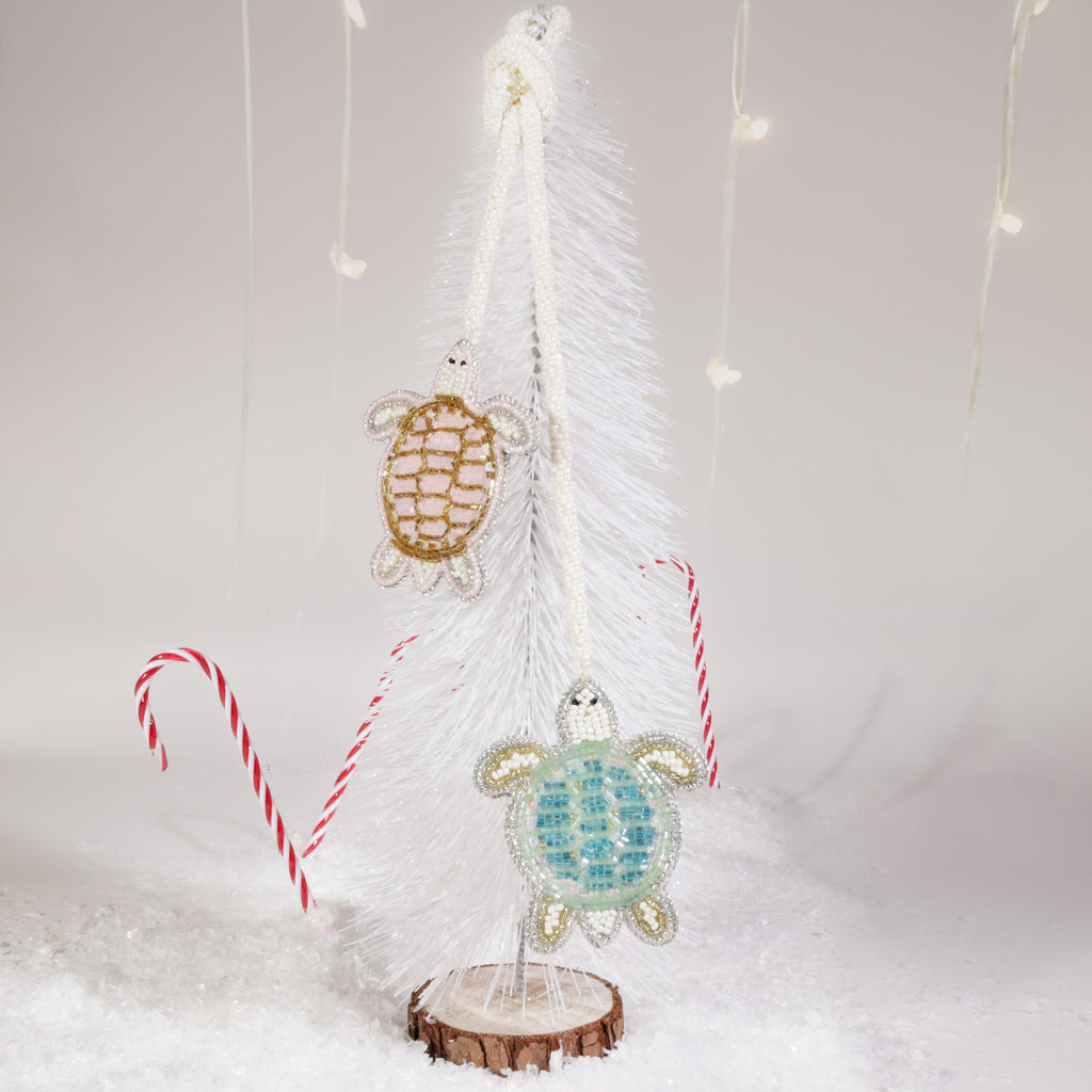 Two beaded turtle bag charms, one gold and one turquoise, hanging from a white decorative tree surrounded by artificial snow, candy canes, and soft fairy lights.