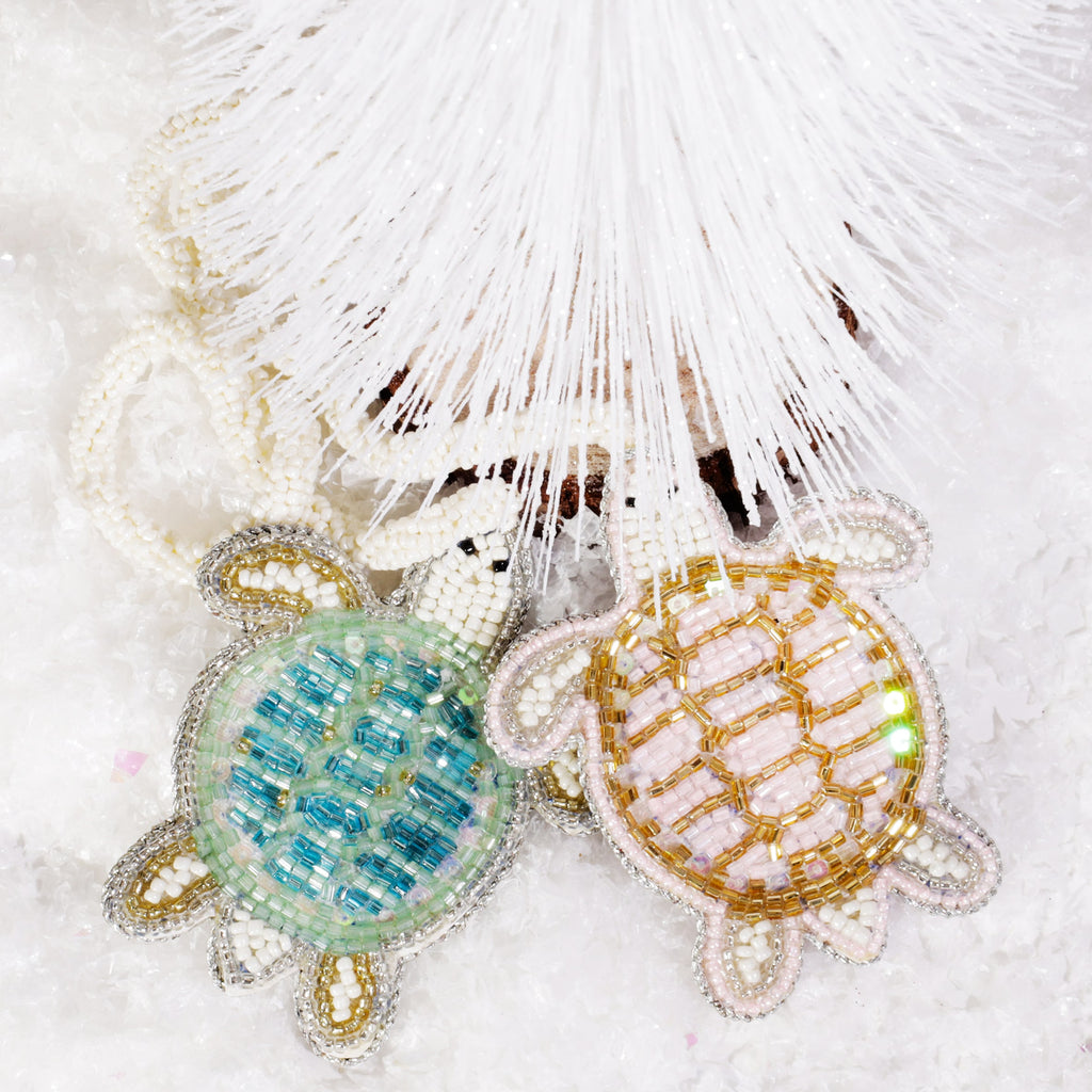 Two beaded turtle bag charms, one turquoise and one gold, lying on artificial snow beneath a white decorative tree.