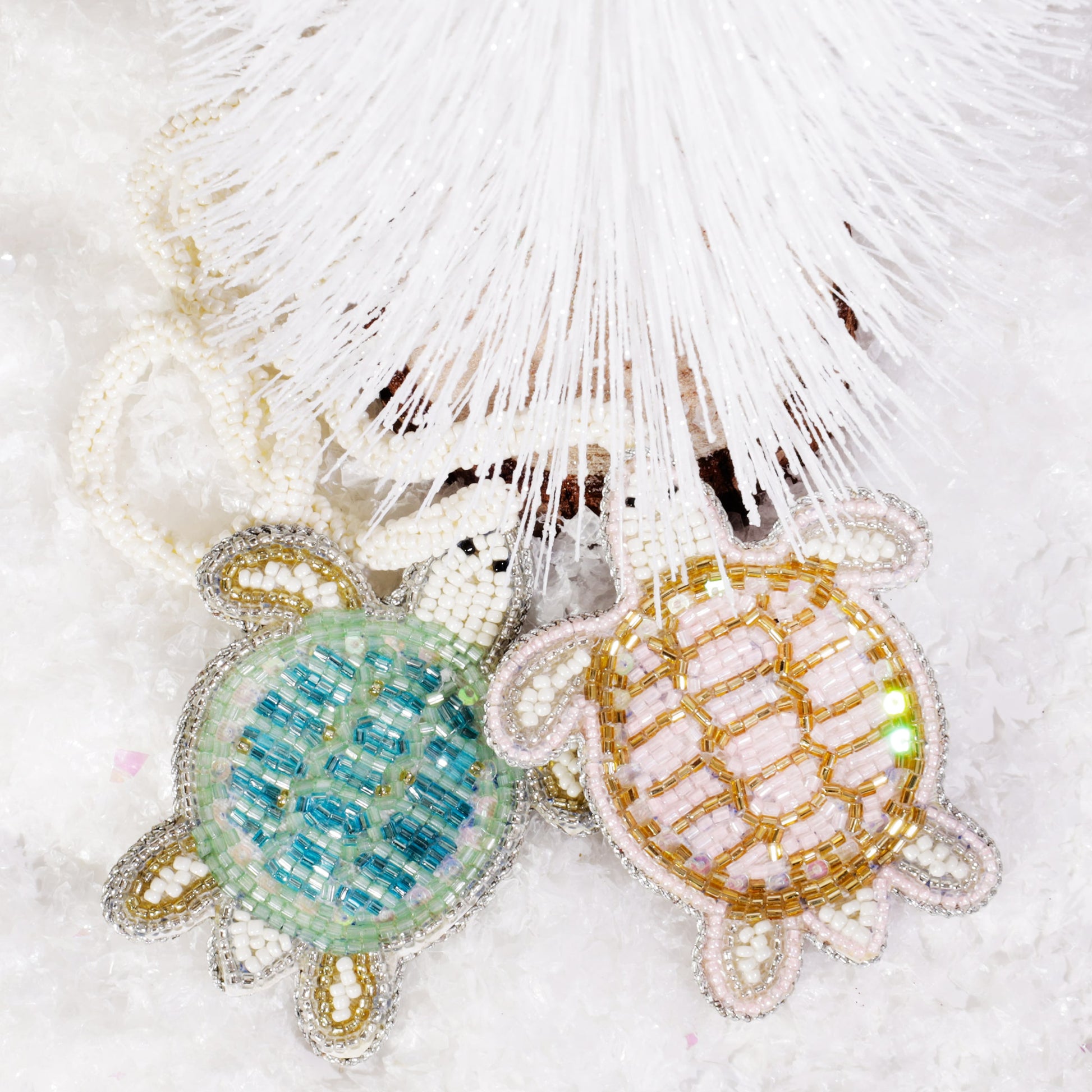Two beaded turtle bag charms, one turquoise and one gold, lying on artificial snow beneath a white decorative tree.