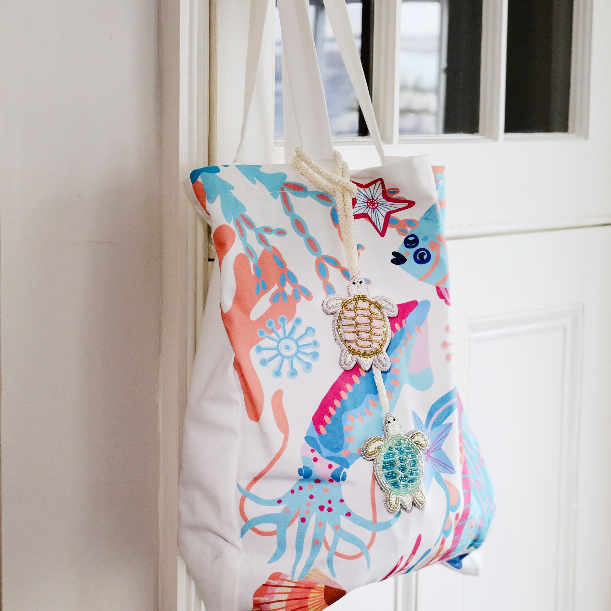 Two beaded turtle bag charms, one gold and one turquoise, hanging from a white tote bag decorated with a colorful coral reef and sea creature print, displayed on a white door.