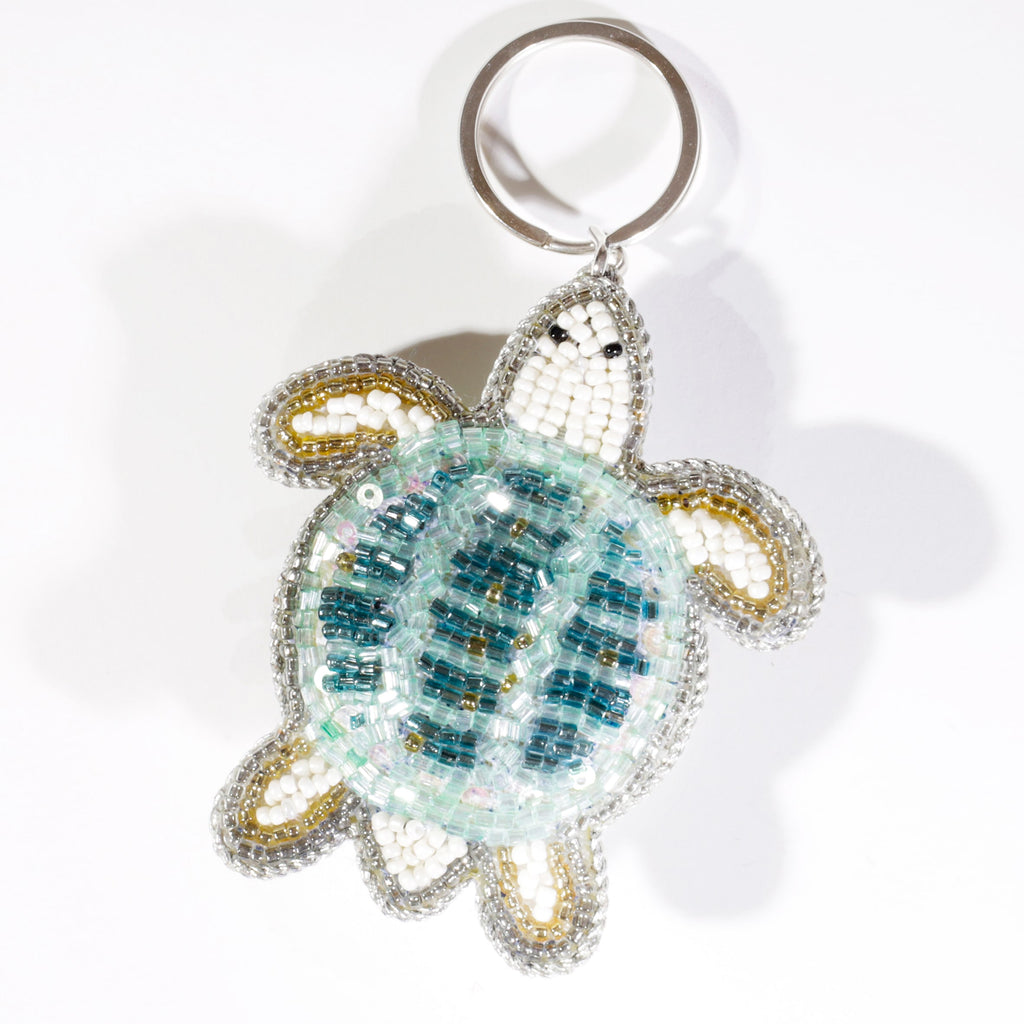 A beaded turtle keyring featuring turquoise, white, silver, and gold beads arranged in a mosaic pattern, displayed against a white background.