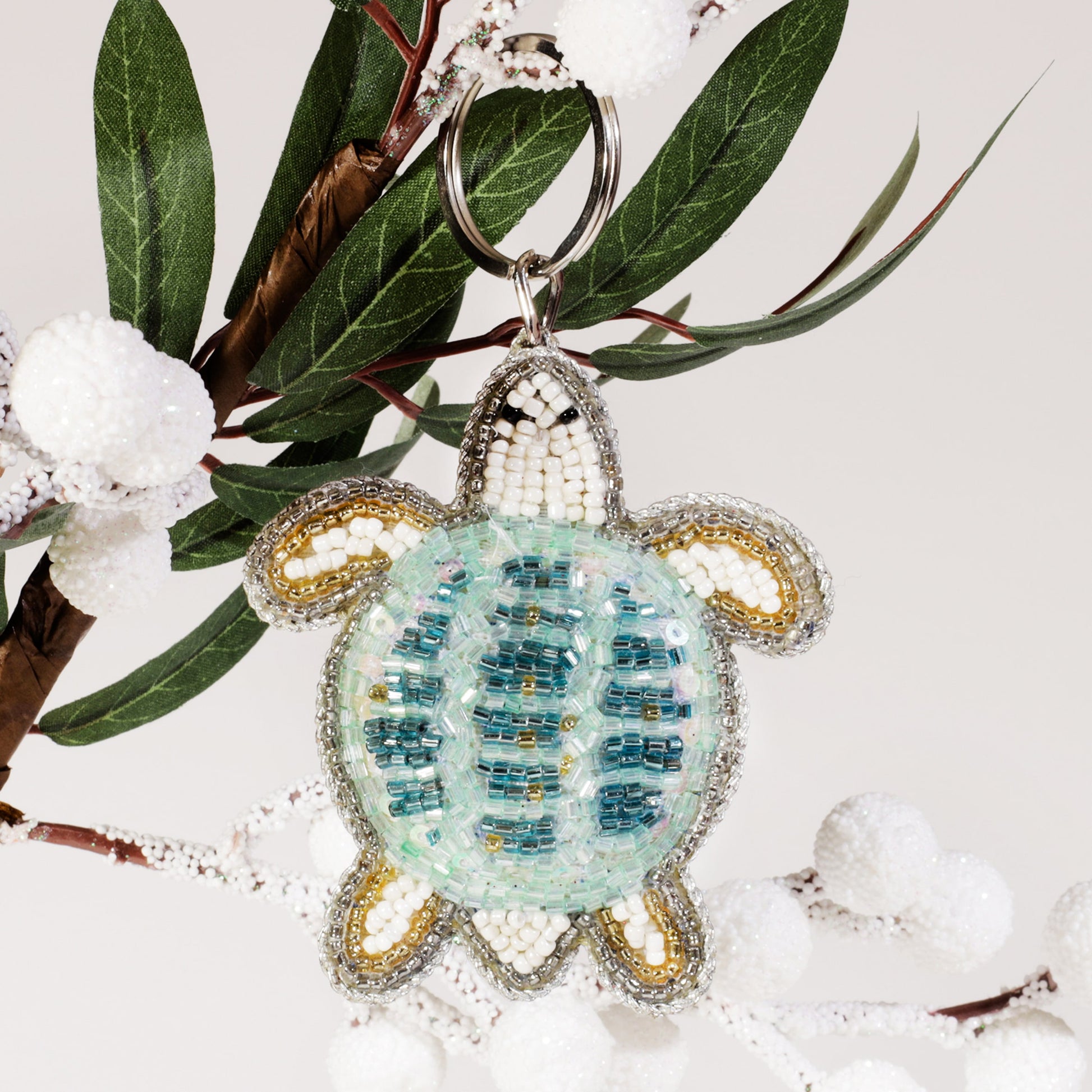 A beaded turtle keyring with turquoise, white, and gold beads hanging from a frosted branch decorated with white berries and green leaves.