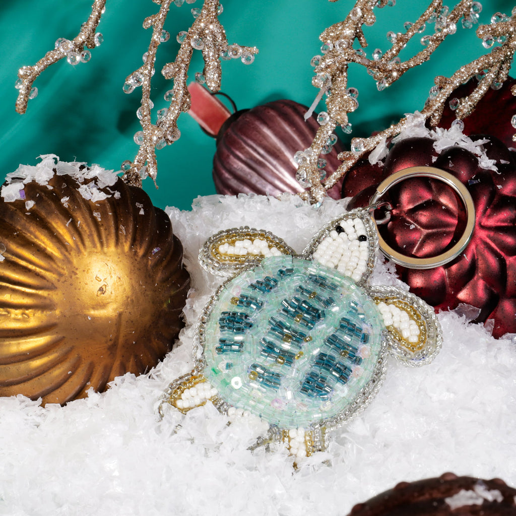 A beaded turtle keyring with turquoise, white, and gold beads resting on artificial snow among red and gold Christmas baubles and glittering gold branches against a teal background.