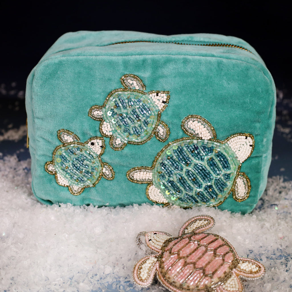 Turquoise velvet wash bag decorated with three beaded sea turtles in shades of blue and gold, displayed on faux snow with a pink beaded turtle decoration in front against a dark festive background.
