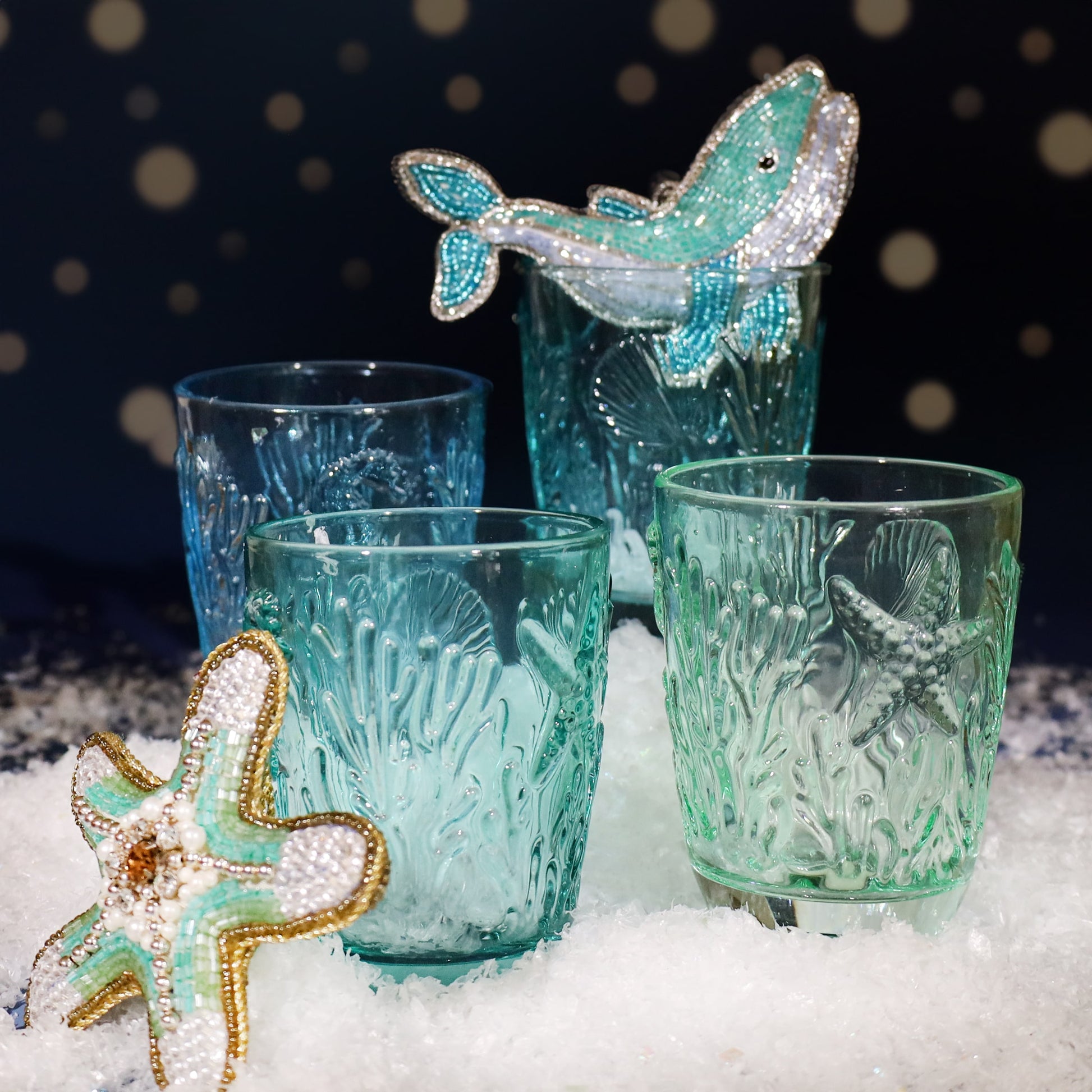 Set of four turquoise glass tumblers with embossed coral and starfish designs, displayed on faux snow with beaded whale and starfish decorations against a softly lit festive background.