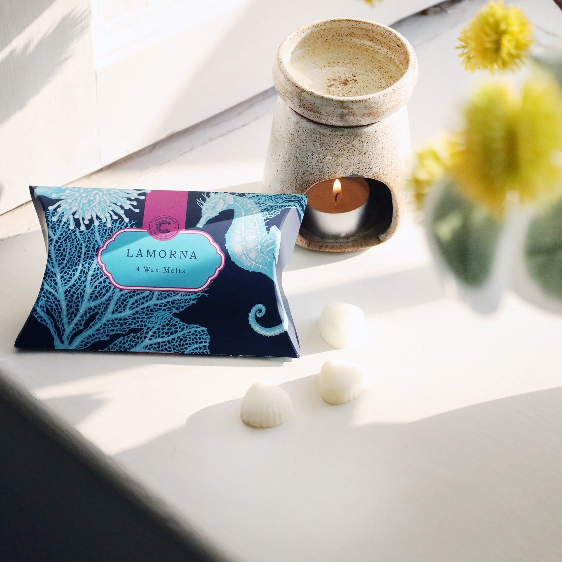 Lamorna wax melts packaging on a white surface with a candle and flowers.