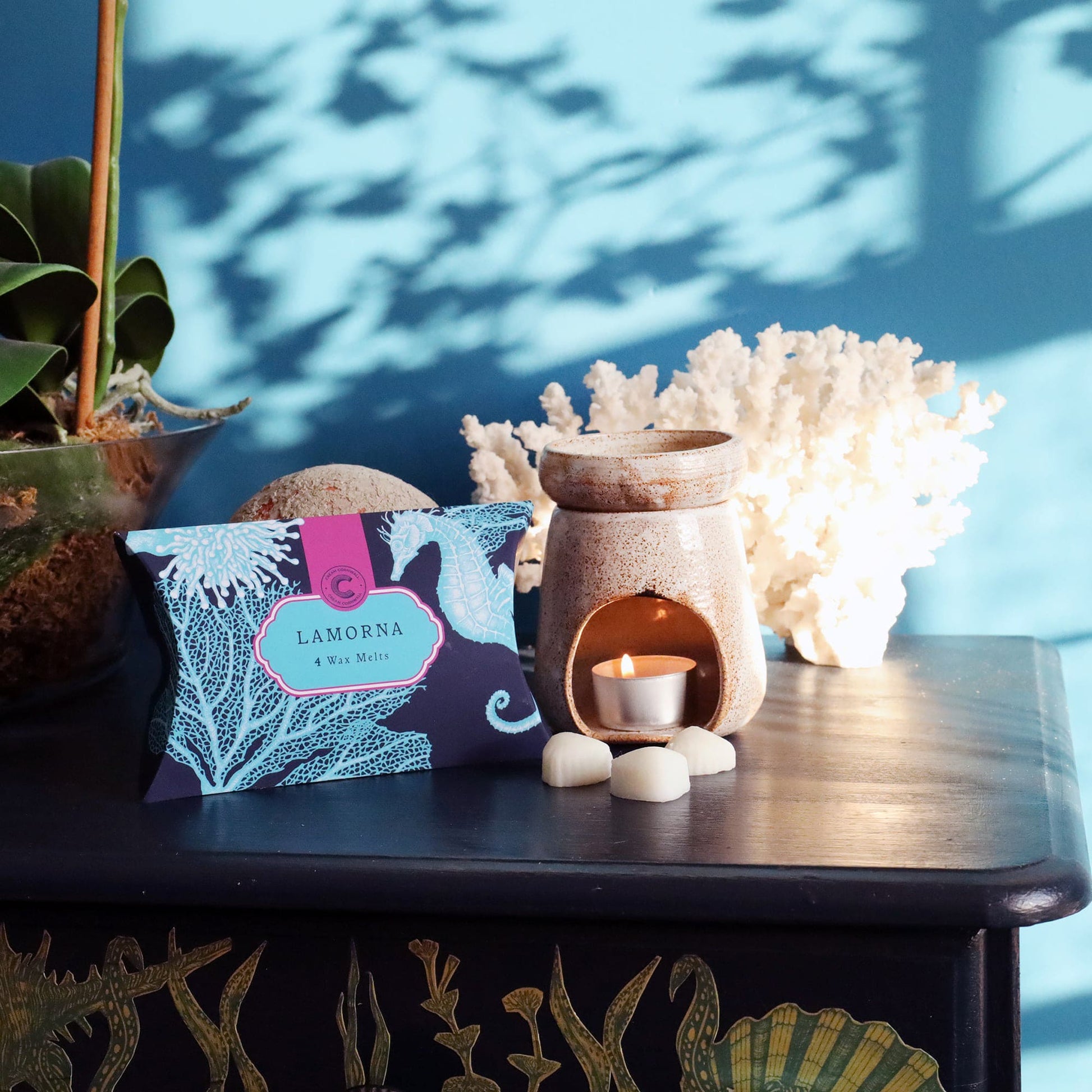 Decorative items including scented Lamorna wax melts, a small plant, and a decorative box on a table with a blue wall in the background.