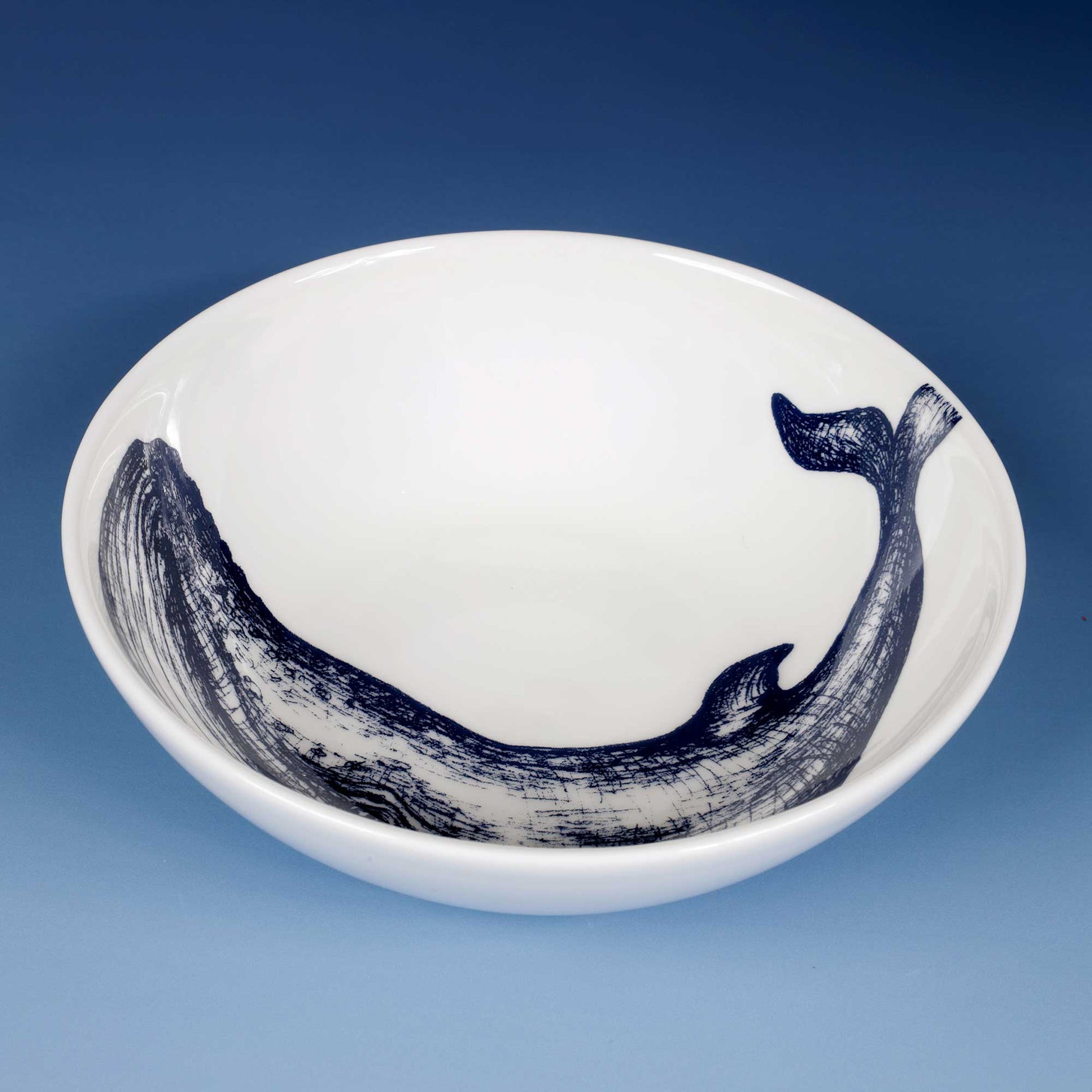 A white ceramic bowl featuring a dark blue whale illustration along the inner rim, set against a blue gradient background.