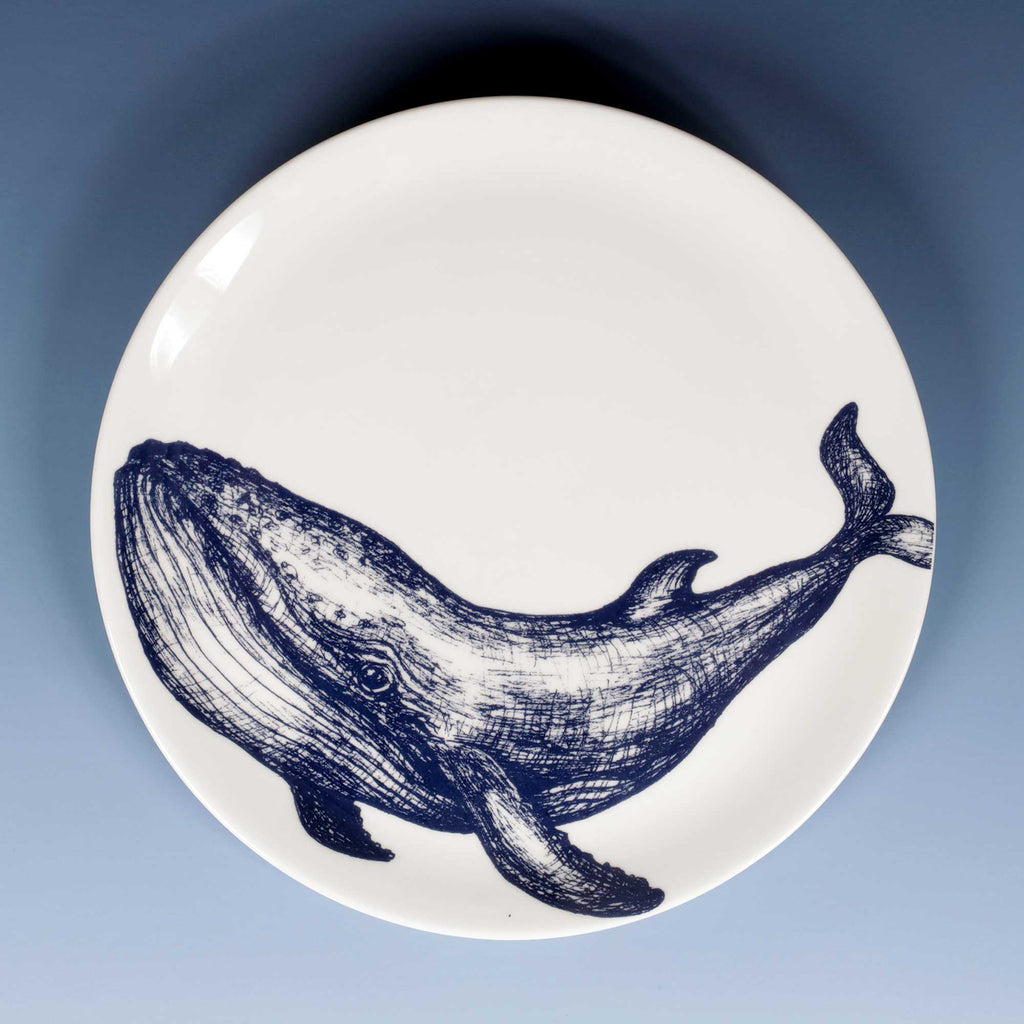 A round white plate featuring a detailed navy blue whale illustration, photographed from directly above against a smooth blue gradient background.
