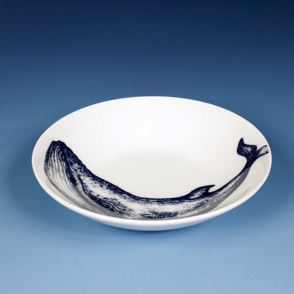 A white ceramic bowl featuring a detailed navy blue whale illustration printed along the inner rim, set against a smooth blue gradient background.