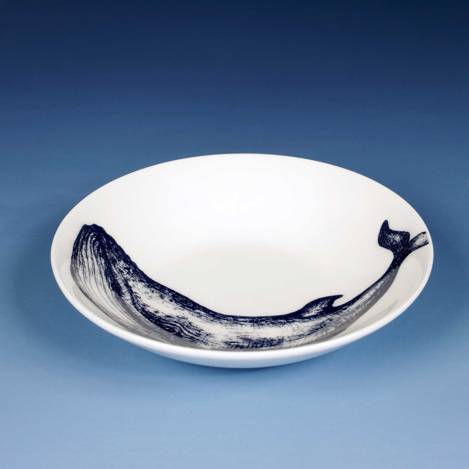A white ceramic bowl featuring a detailed navy blue whale illustration printed along the inner rim, set against a smooth blue gradient background.