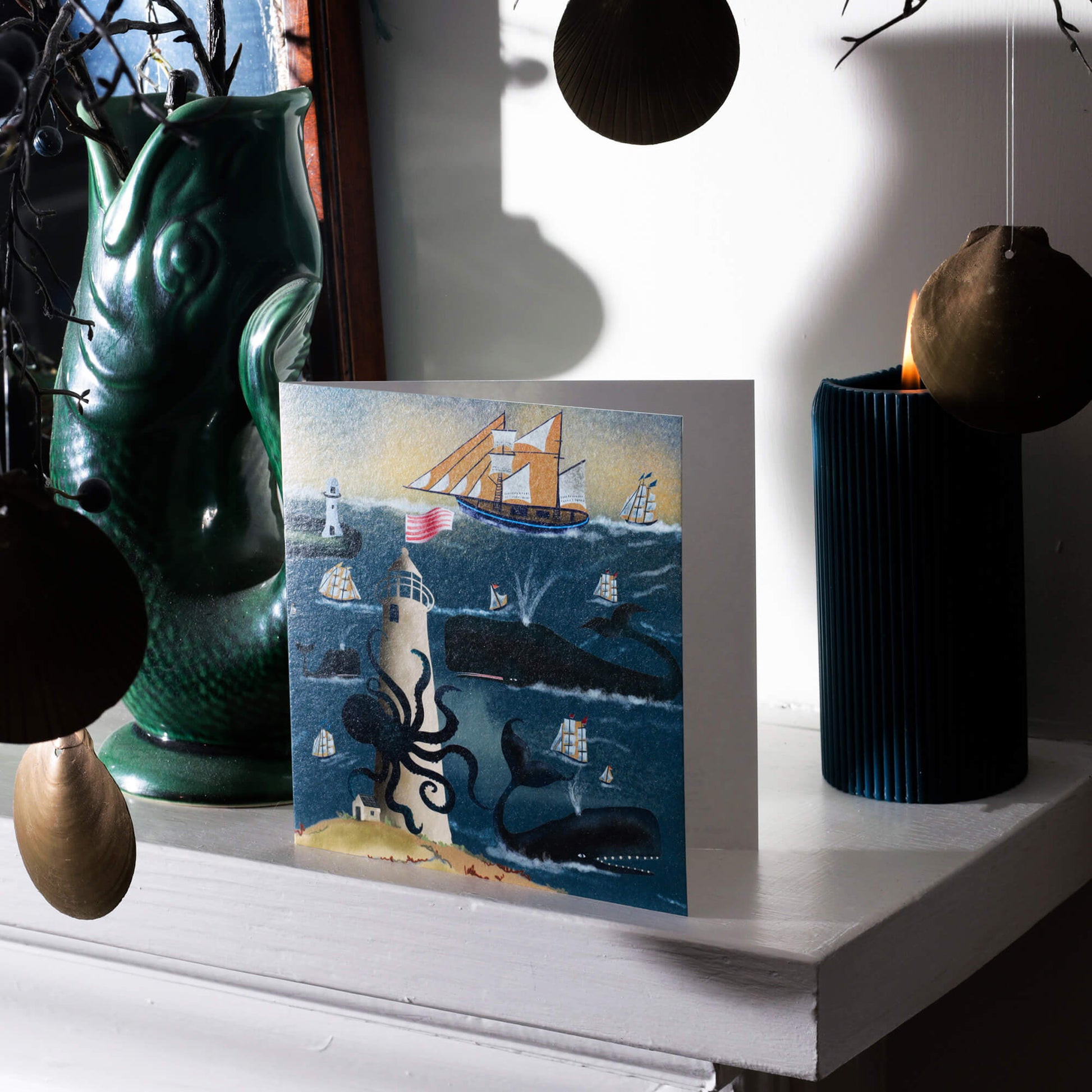 A nautical-themed greeting card featuring whales, ships, and a lighthouse, displayed on a mantelpiece. The card is surrounded by coastal decor, including hanging seashell ornaments, a dark green ceramic vase, and a lit blue candle. The scene is bathed in soft natural light, creating a calm, seaside-inspired atmosphere.