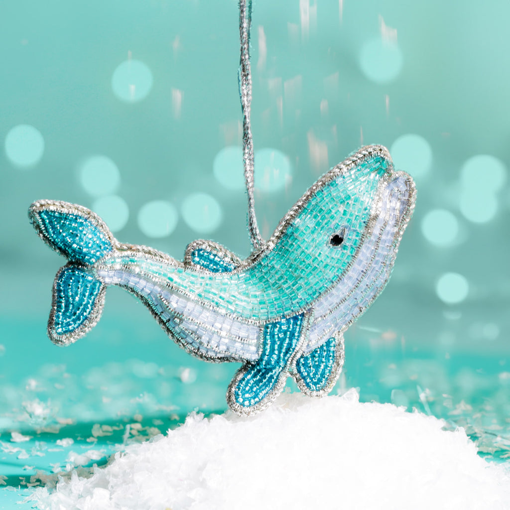 A turquoise and white beaded whale decoration with silver trim, hanging above artificial snow against a teal background with softly glowing lights.