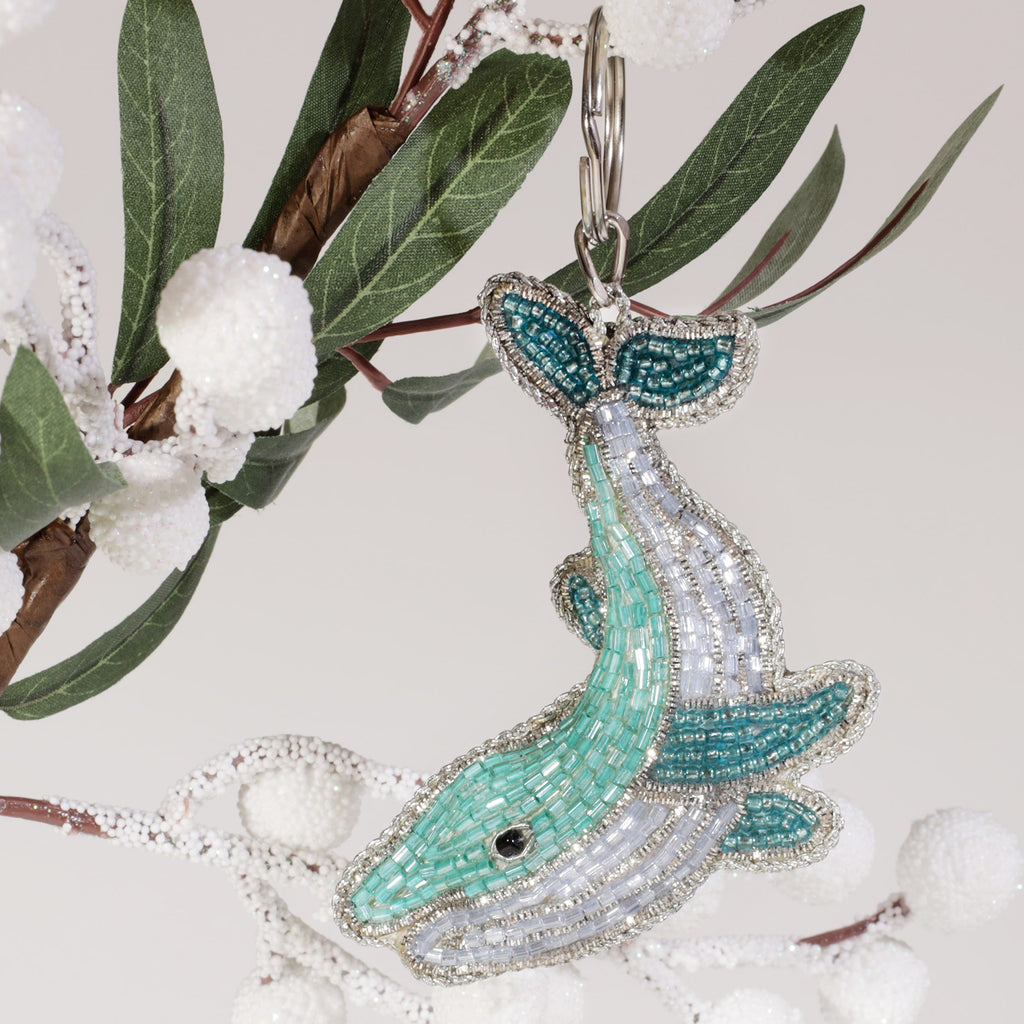 A turquoise and silver beaded whale keyring with intricate detailing, hanging from a branch decorated with white frosted berries and green leaves.