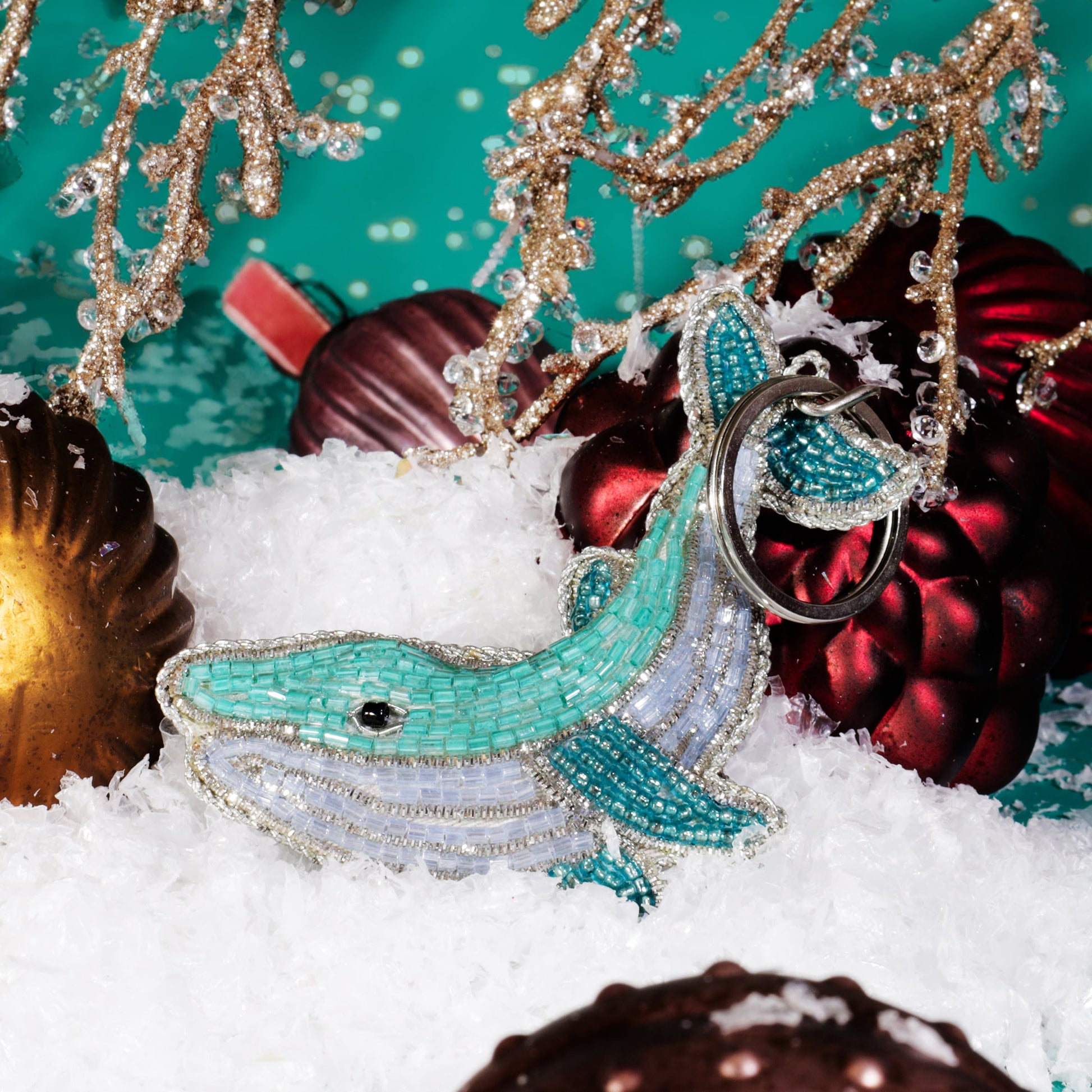 A turquoise and silver beaded whale keyring displayed on artificial snow among red and gold Christmas baubles and glittering gold branches against a teal background.